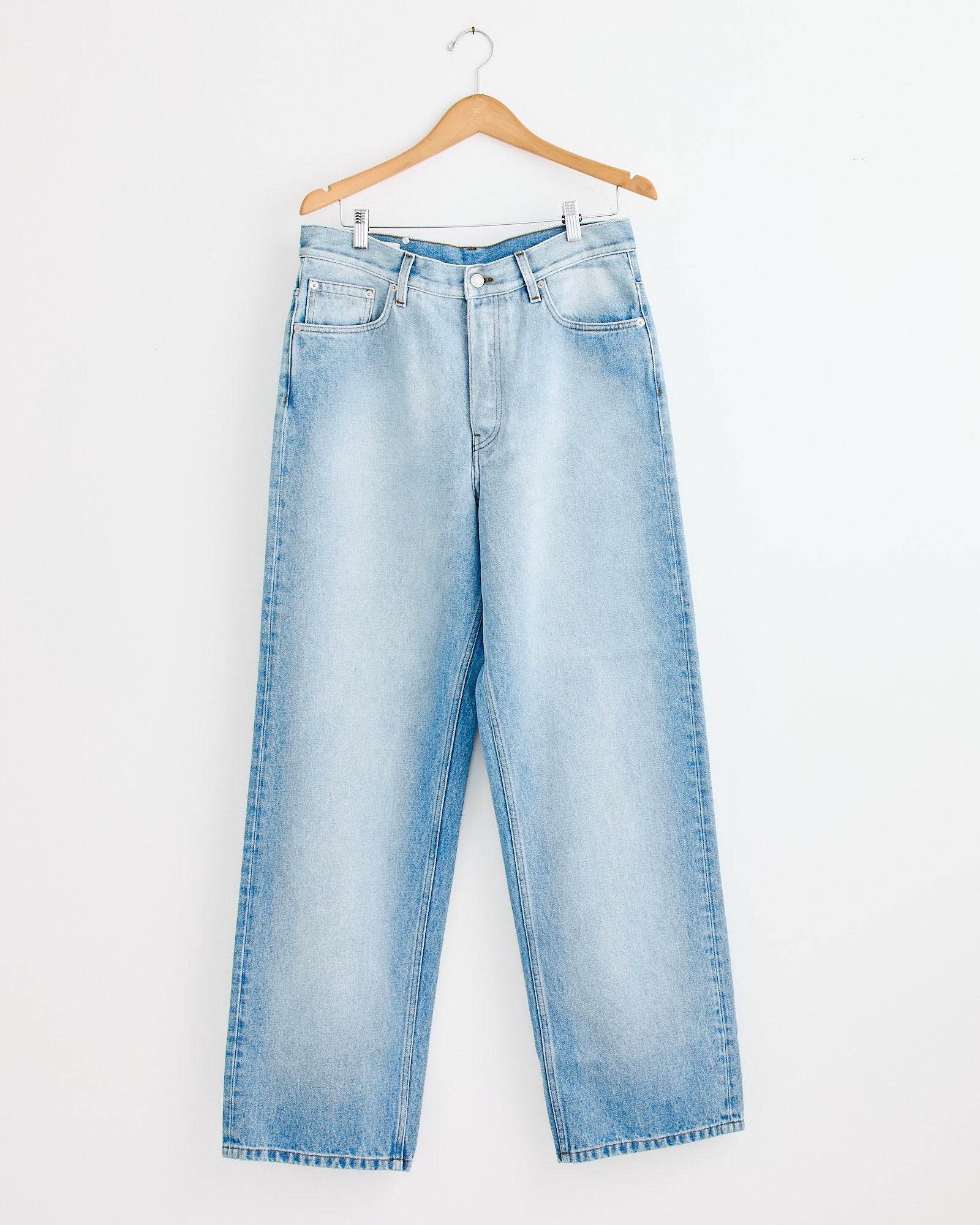 The Pine Den 2390 Pant in Light Blue by Dries Van Noten features wide legs, a high waist, five pockets, and a button fly. Shown on a wooden hanger against a white wall. For sizing help, contact our online chat.