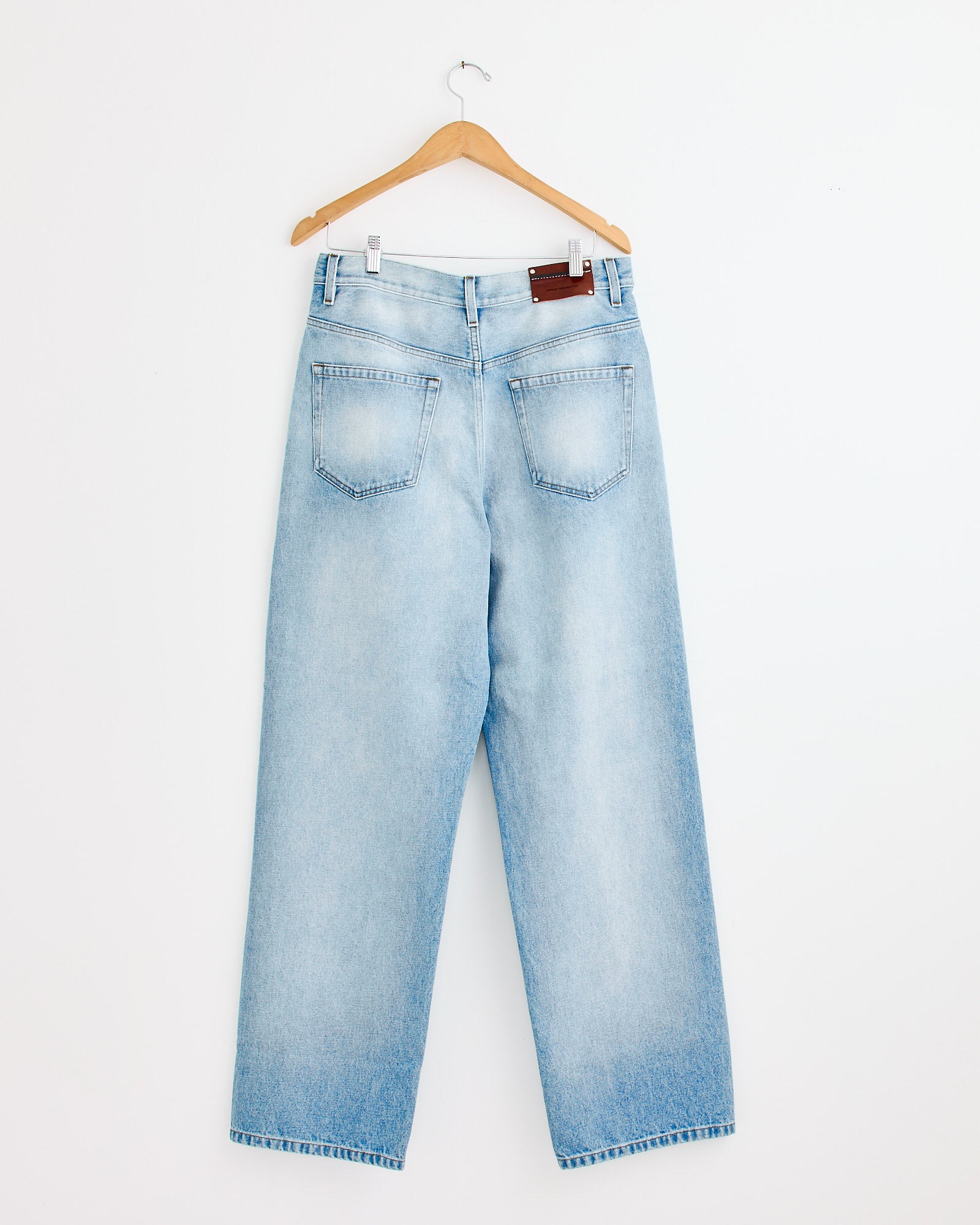 The Dries Van Noten Pine Den 2390 Pant in Light Blue—high-waisted, straight-leg jeans with back pockets—hangs on a wooden hanger against a white wall. For sizing help, contact our online chat team for personalized assistance.