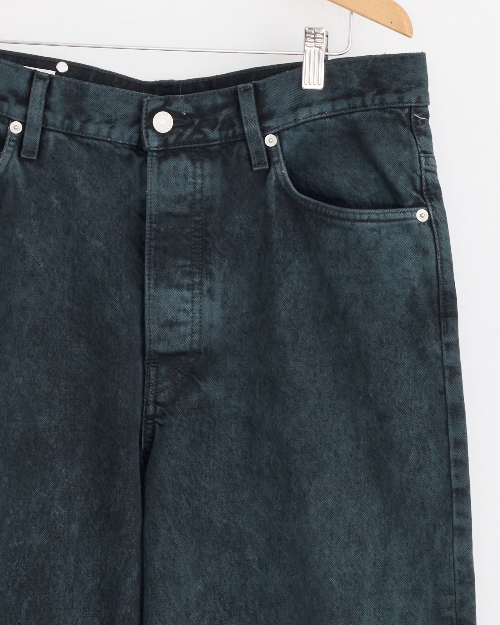 Close-up of Dries Van Noten Loose Fit Jeans in Petrol hanging on a wooden hanger against a white background, showcasing heavyweight denim, waistband, pockets, and front button details.