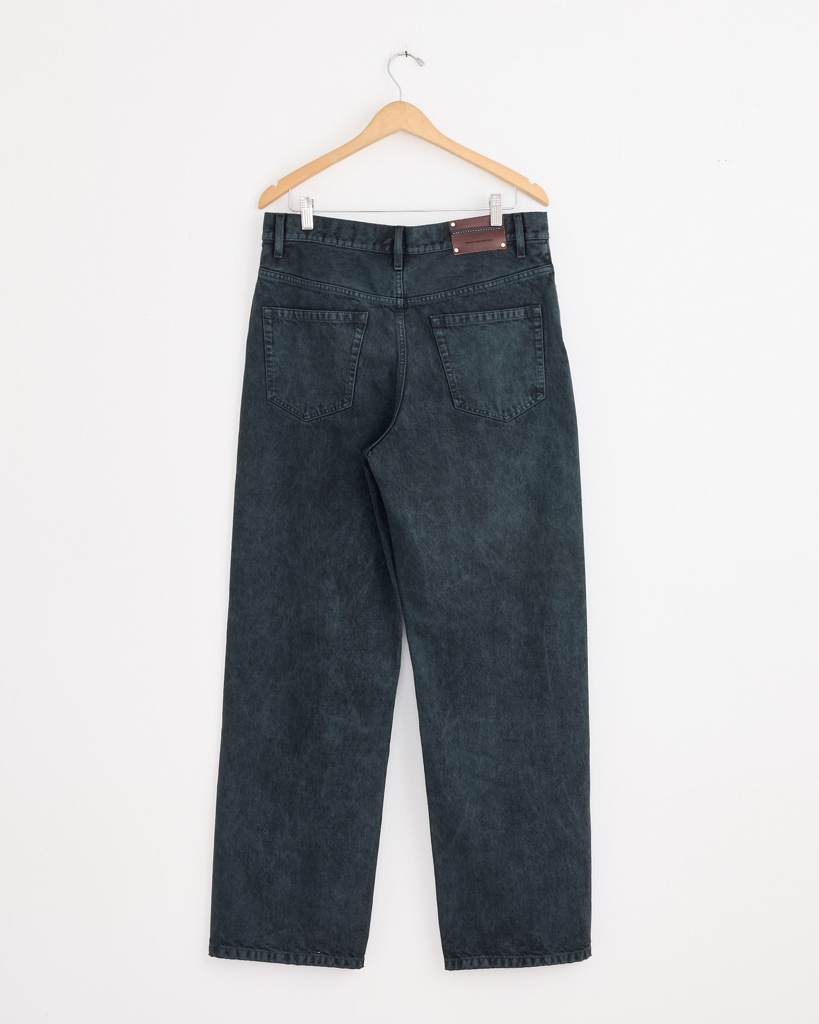 A pair of Dries Van Noten Loose Fit Jeans in Petrol hangs on a wooden hanger against a plain white wall, displayed with the back side forward and two visible back pockets.