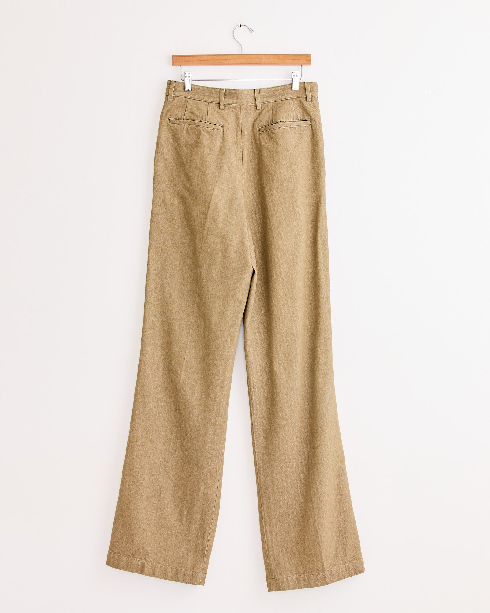 The Dries Van Noten Pleated Denim Pant in Stone, featuring back pockets and a tailored wide-leg fit, hangs on a wooden hanger against a plain white wall.