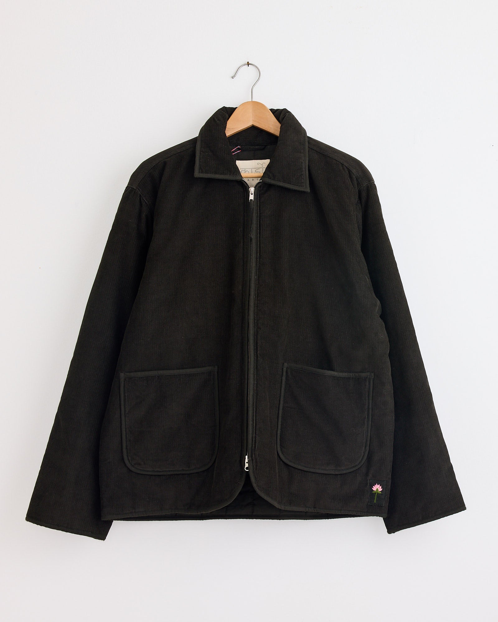 The Kardo Amardeep Jacket in Dark Green features a collar, front zipper, and two large front pockets. Displayed on a wooden hanger against a white wall. For fit or sizing questions, contact us via our online chat.