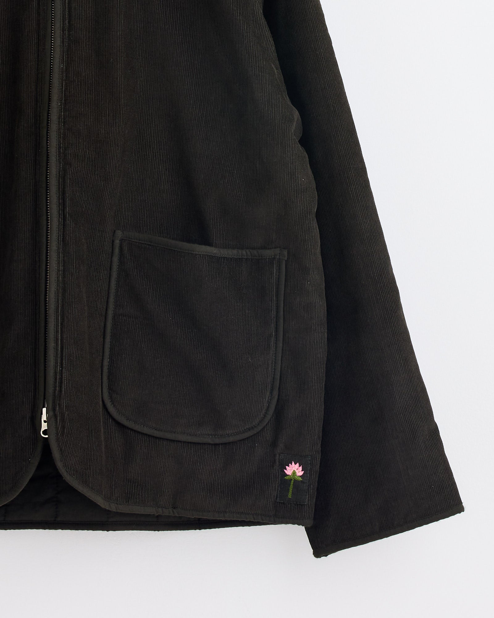 A close-up of the Kardo Amardeep Jacket in Dark Green, featuring a large front pocket and a small embroidered pink flower near the hem. The partially unzipped corduroy jacket is displayed against a plain white background—perfect for any fit.