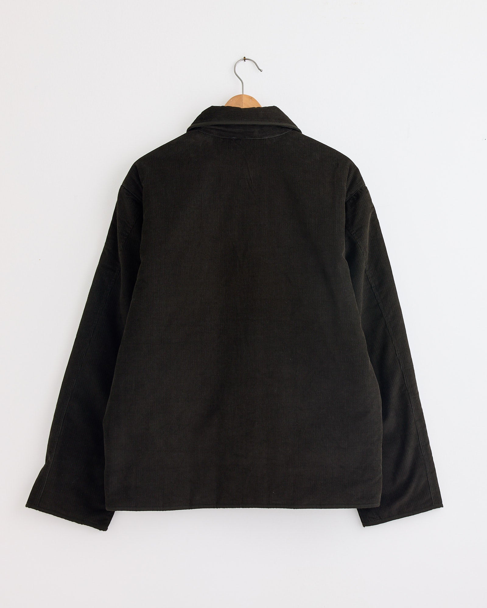The Amardeep Jacket in Dark Green by Kardo, a corduroy style with long sleeves, hangs on a wooden hanger against a white wall shown from the back. For fit or sizing questions, connect with our online chat for assistance.