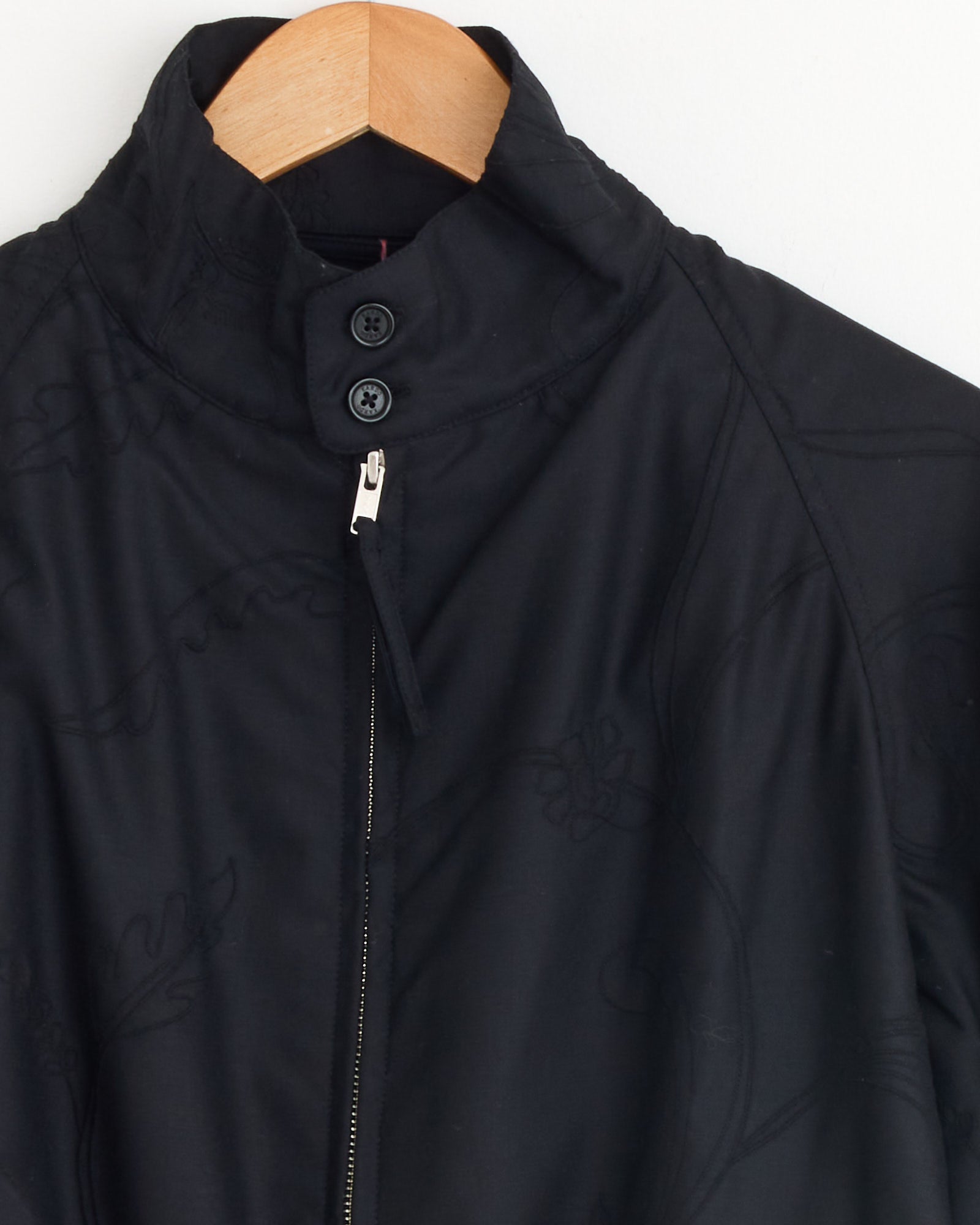 A close-up of the Kardo Stanley Jacket in Black, styled like a Harrington with a high collar, two black buttons, a zipper, and subtle stitched patterns, hanging on a wooden hanger against a white background.