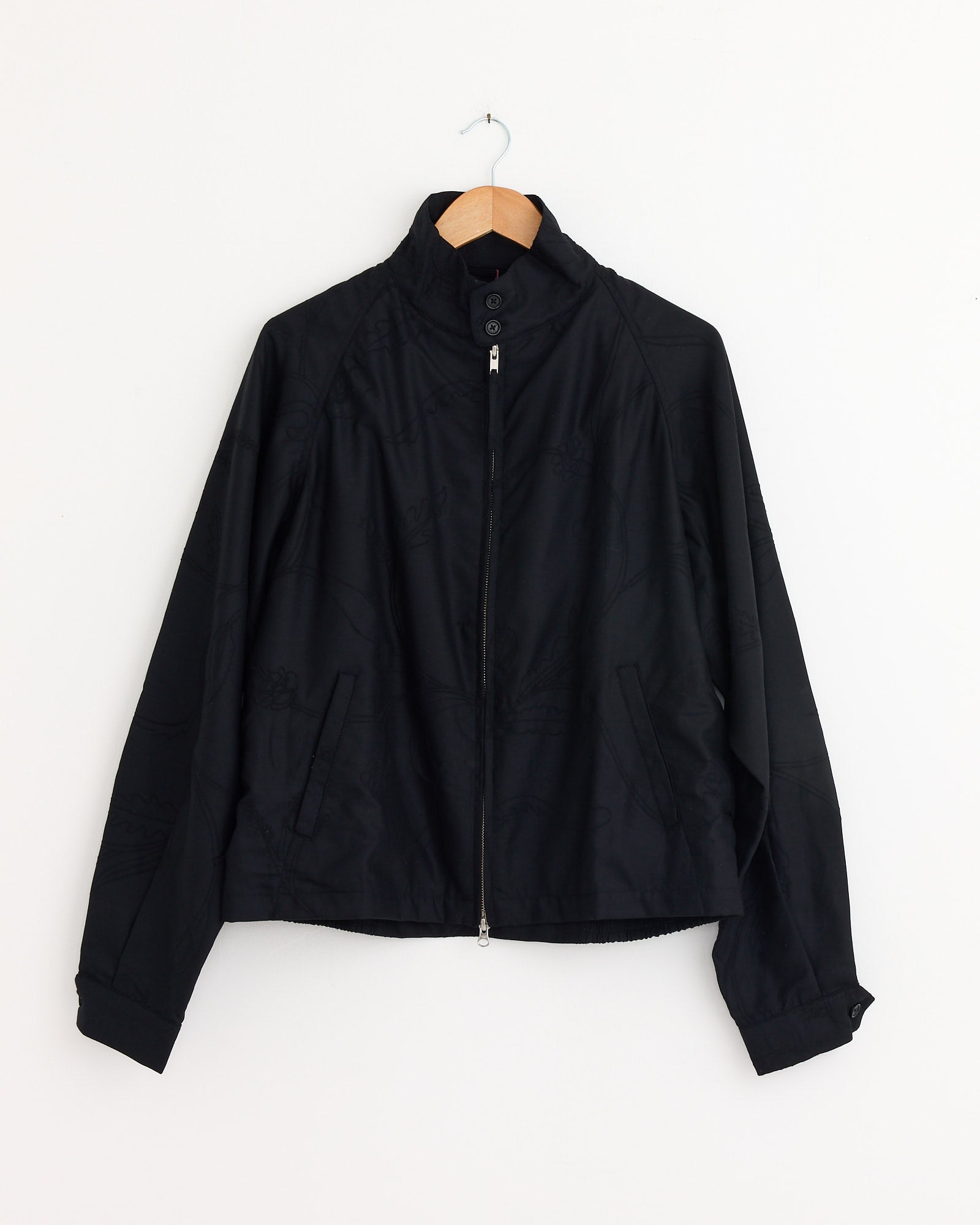 The Kardo Stanley Jacket in Black, crafted from Super 120's wool, hangs on a wooden hanger by a white wall. Designed in a classic Harrington style, it features a high collar, long sleeves, and two slanted front pockets.