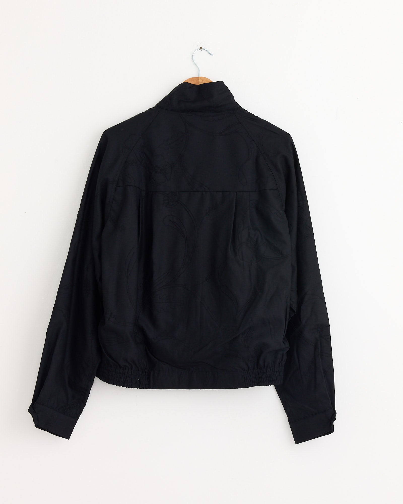 The Kardo Stanley Jacket in Black hangs on a wooden hanger against a plain white wall, shown from the back with long sleeves and an elastic waistband.