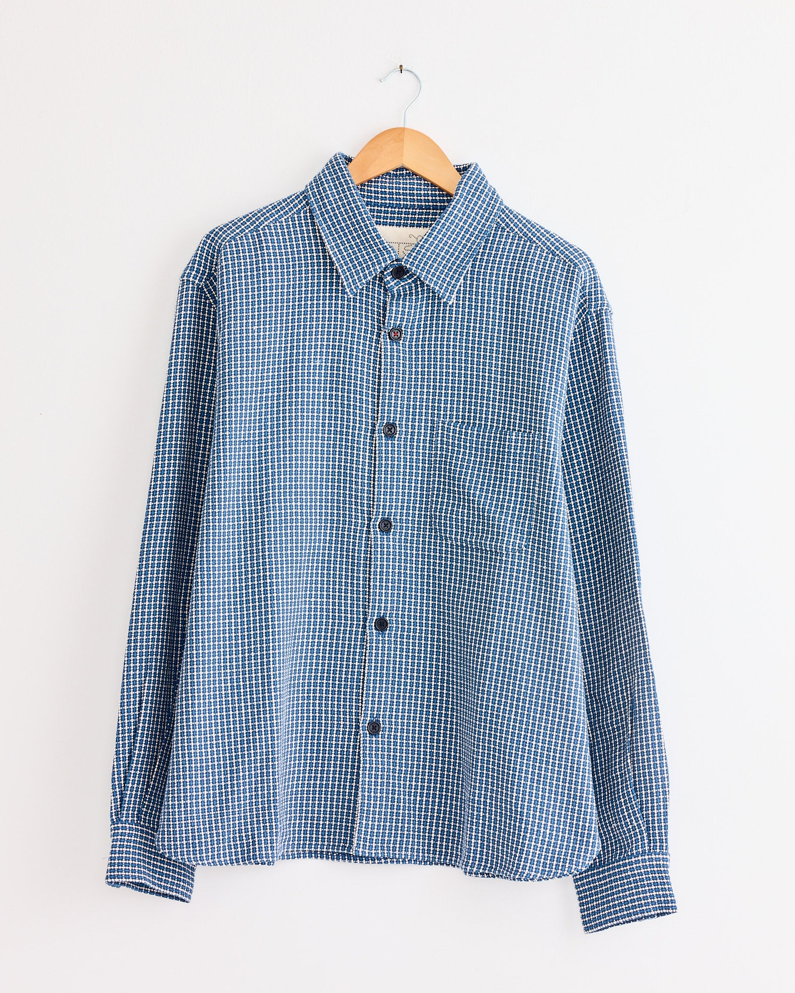 The Kardo Luis Shirt in Indigo, featuring a blue and white checkered pattern, hangs on a wooden hanger against a plain white wall.