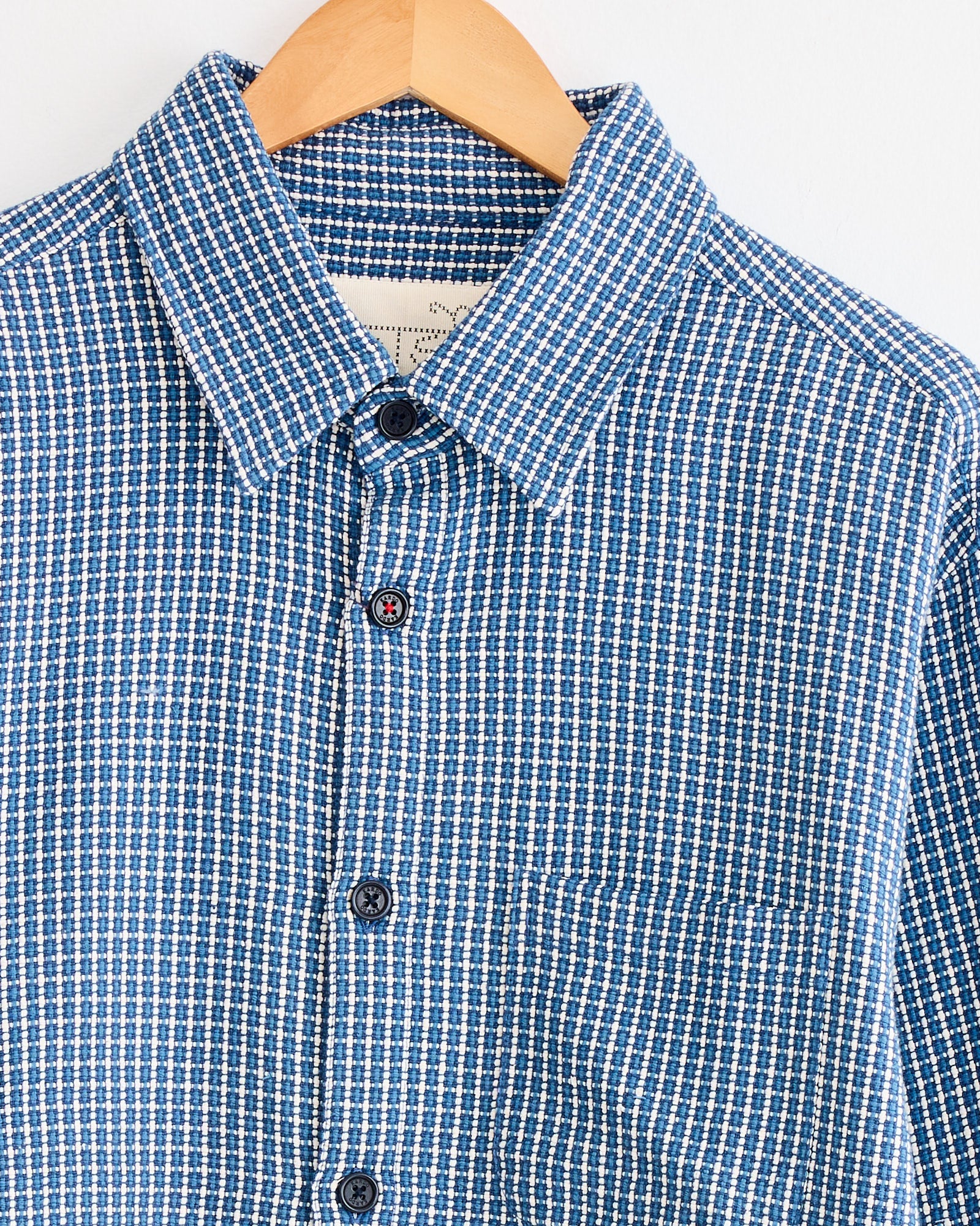 The Luis Shirt in Indigo by Kardo, a blue and white checkered khadi cotton shirt with a front pocket, is displayed on a wooden hanger against a white background.