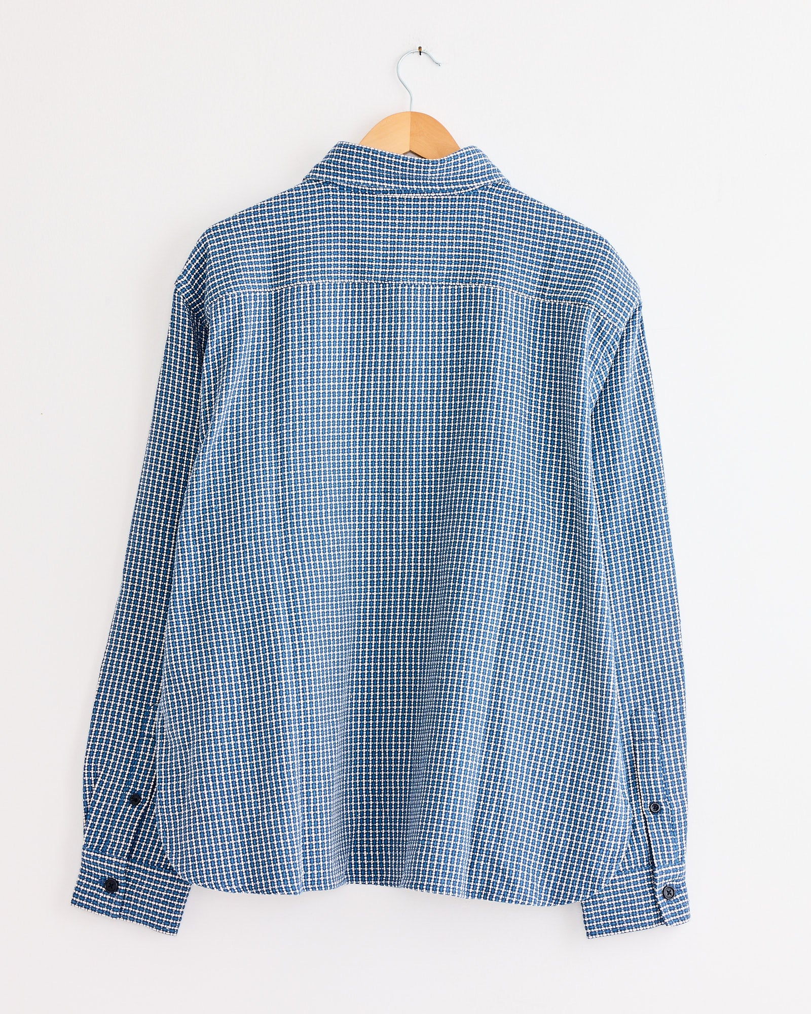 The Luis Shirt in Indigo by Kardo, featuring blue and white checks, hangs on a wooden hanger against a plain white wall, shown from the back.