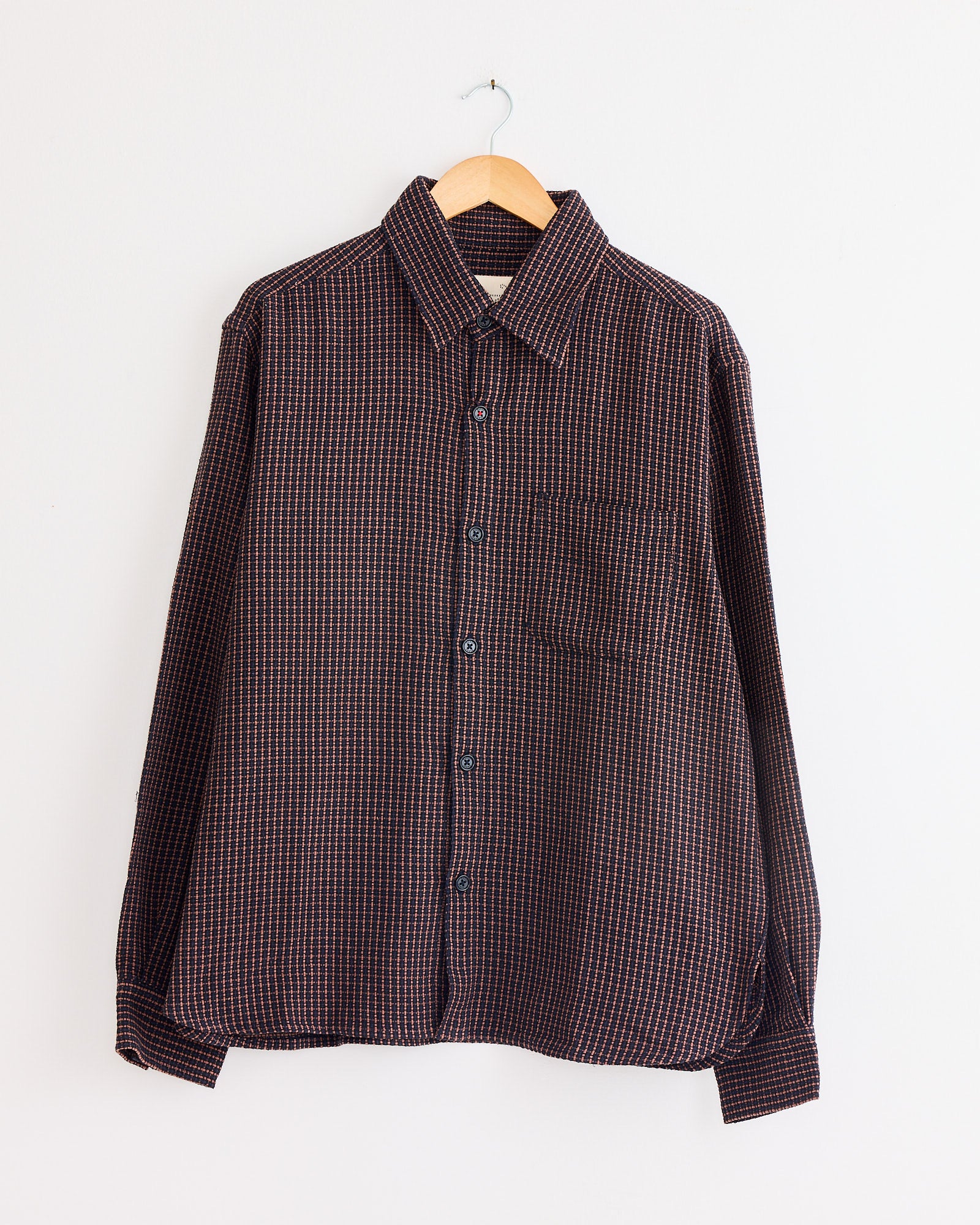 The Kardo Luis Shirt in Ganache, featuring a dark small checkered pattern and long sleeves, is displayed on a wooden hanger against a plain white wall.