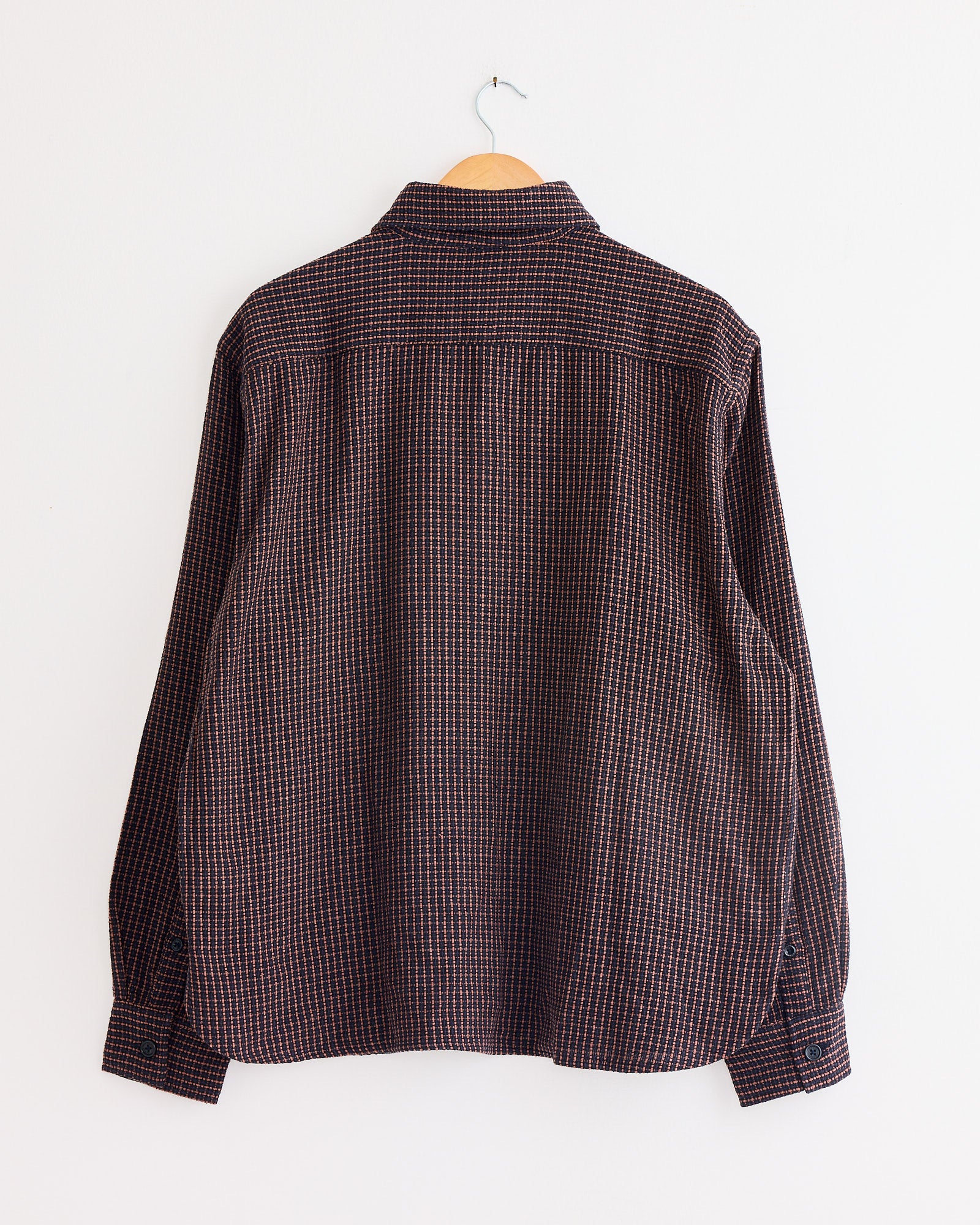 The Luis Shirt in Ganache by Kardo, a long-sleeved dark plaid shirt with red and blue checks, hangs on a wooden hanger against a plain white wall, shown from the back.
