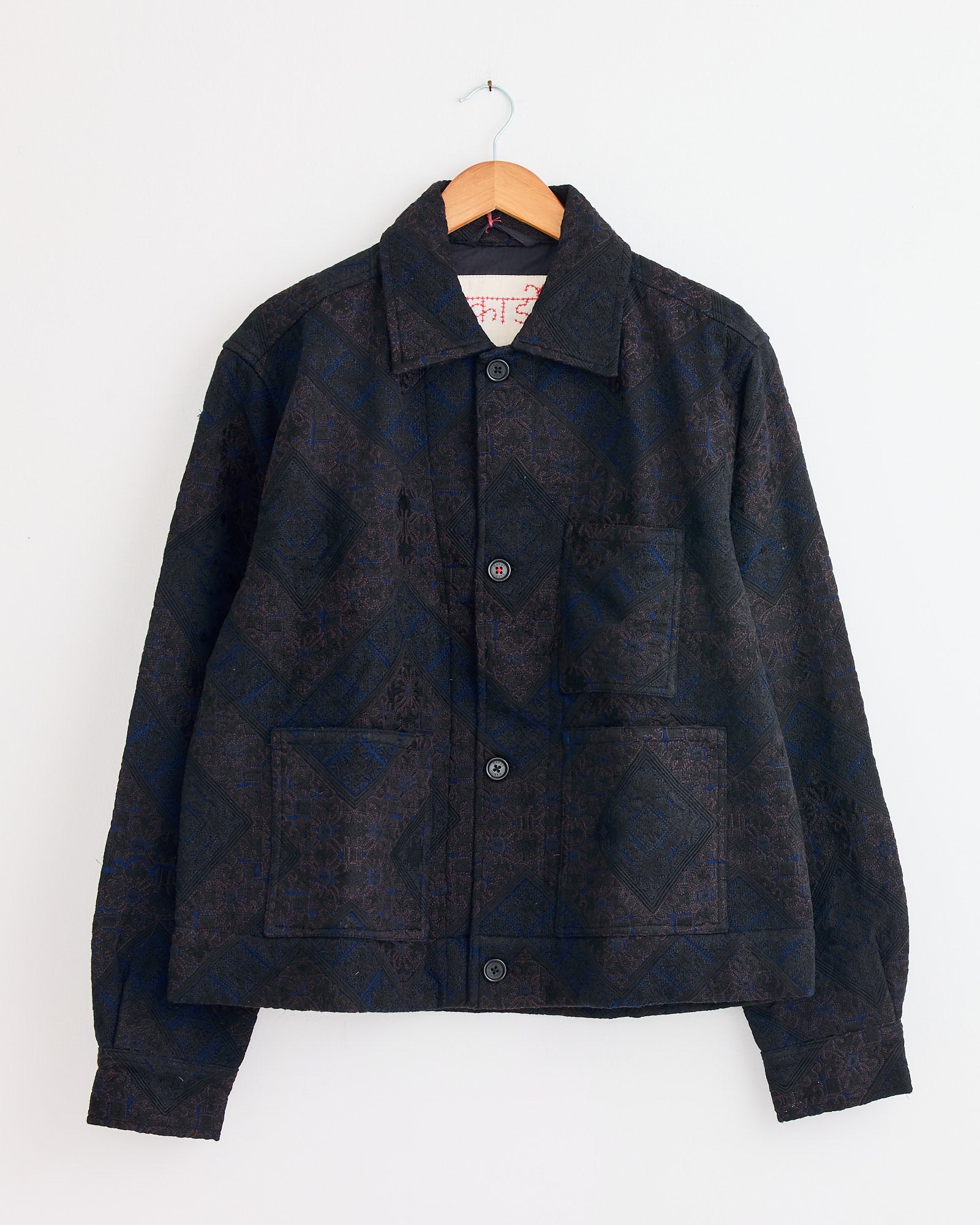 The Bodhi Jacket in Indigo by Kardo features a dark pattern, pointed collar, long sleeves, and two large front pockets. Shown hanging on a wooden hanger against a white wall. Questions about sizing or fit? Contact us via online chat.