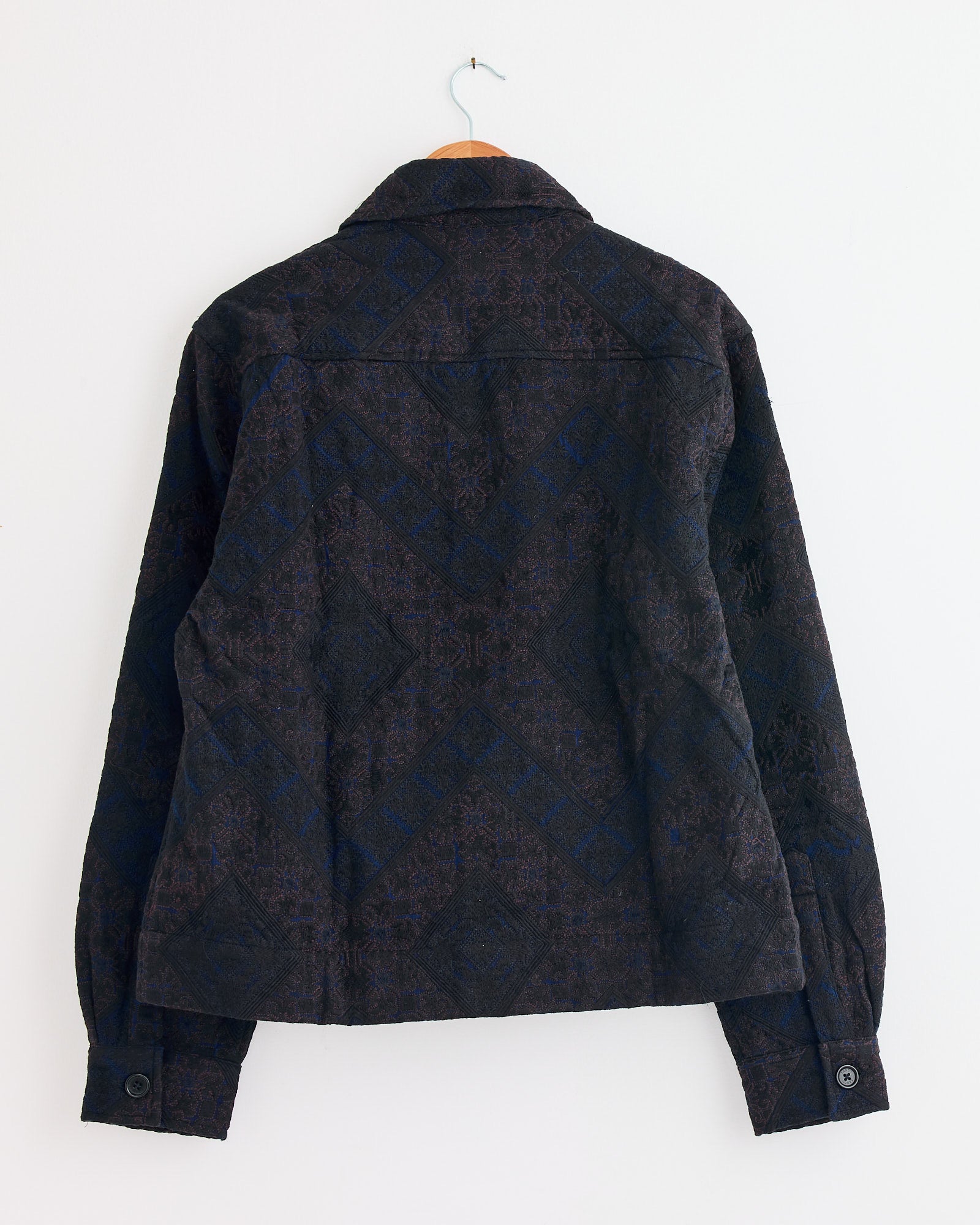 The Bodhi Jacket in Indigo by Kardo is displayed from the back on a wooden hanger against a white wall, highlighting its patterned fabric, collar, long sleeves, and buttoned cuffs—ideal for viewing fit and sizing details.