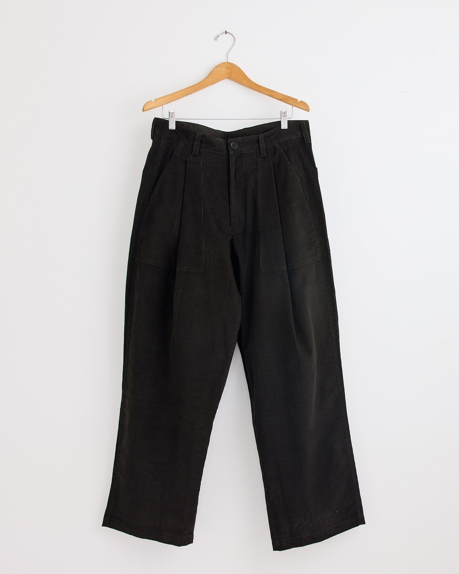 The Kardo Yuki Trouser in Dark Green, featuring wide legs, pleats, front pockets, and a button closure, hangs on a wooden hanger against a white wall. For sizing or fit questions, our online chat is here to help.