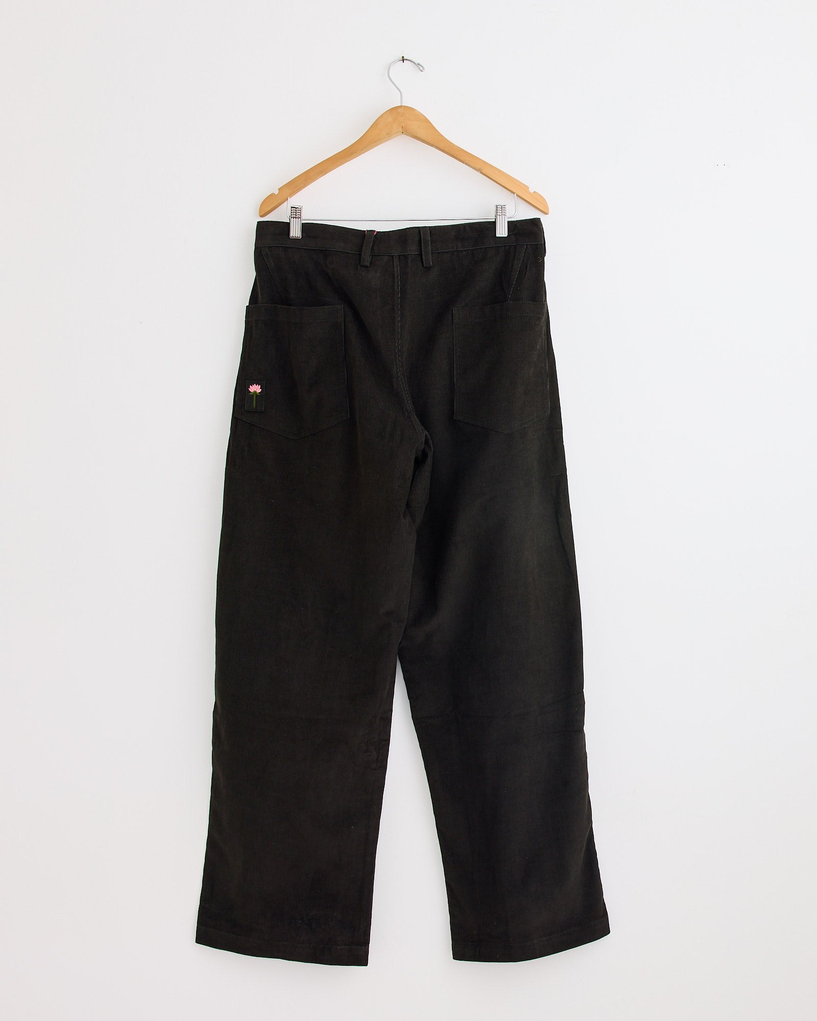 The Kardo Yuki Trouser in Dark Green, featuring a loose fit and two large back pockets (one with a small embroidered design), hangs on a wooden hanger against a plain white wall. For fit or sizing questions, contact us via online chat.