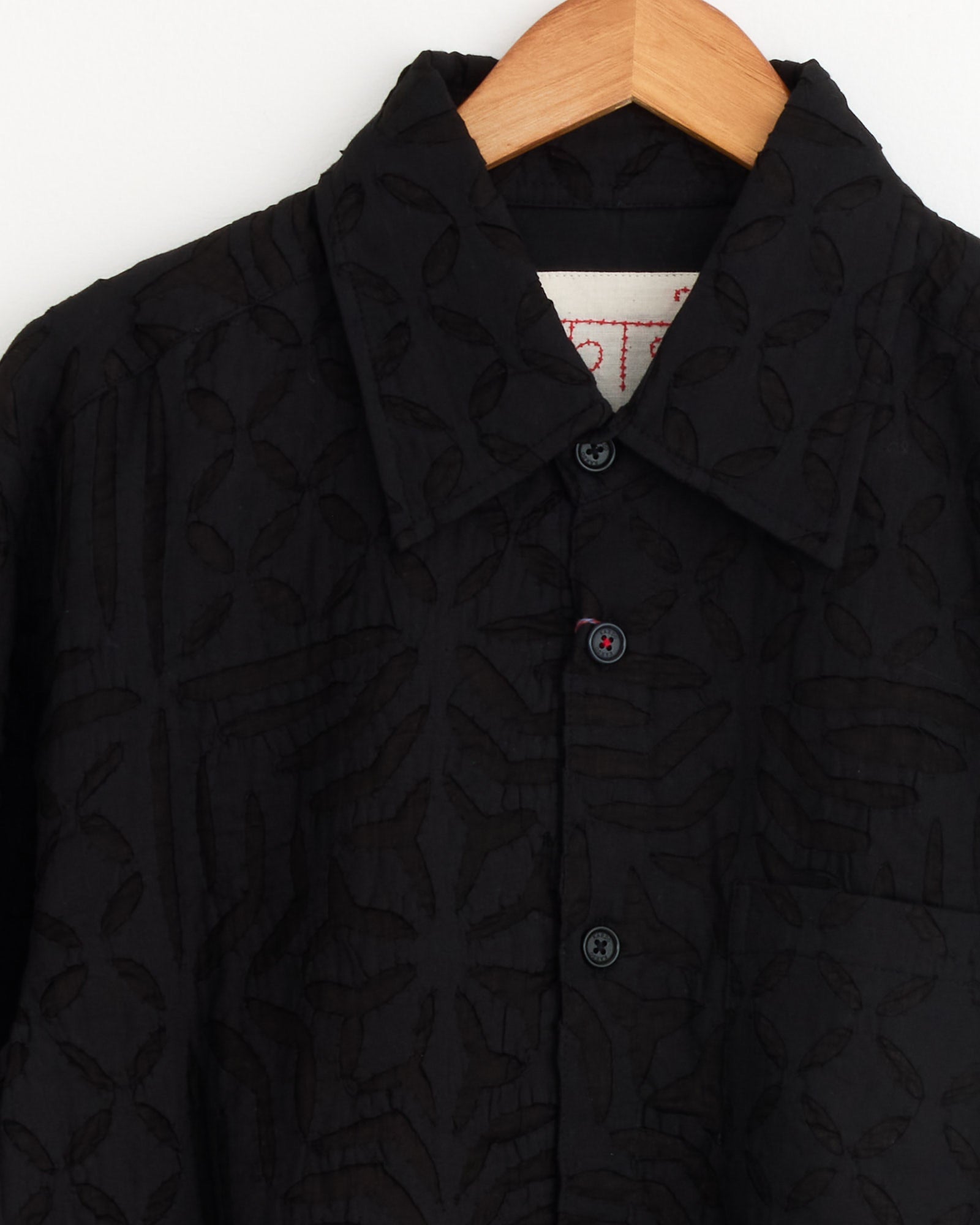 A close-up of the Luis Shirt in Black by Kardo, crafted from black khadi cotton with a collar, subtle leaf pattern, black buttons, and displayed on a wooden hanger—an eco-friendly shirt choice.