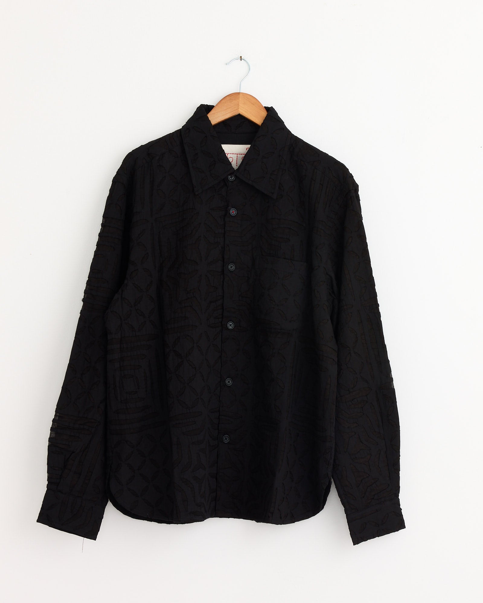 The Kardo Luis Shirt in Black, a long-sleeved khadi cotton shirt with a subtle textured geometric pattern, hangs on a wooden hanger against a plain white wall.