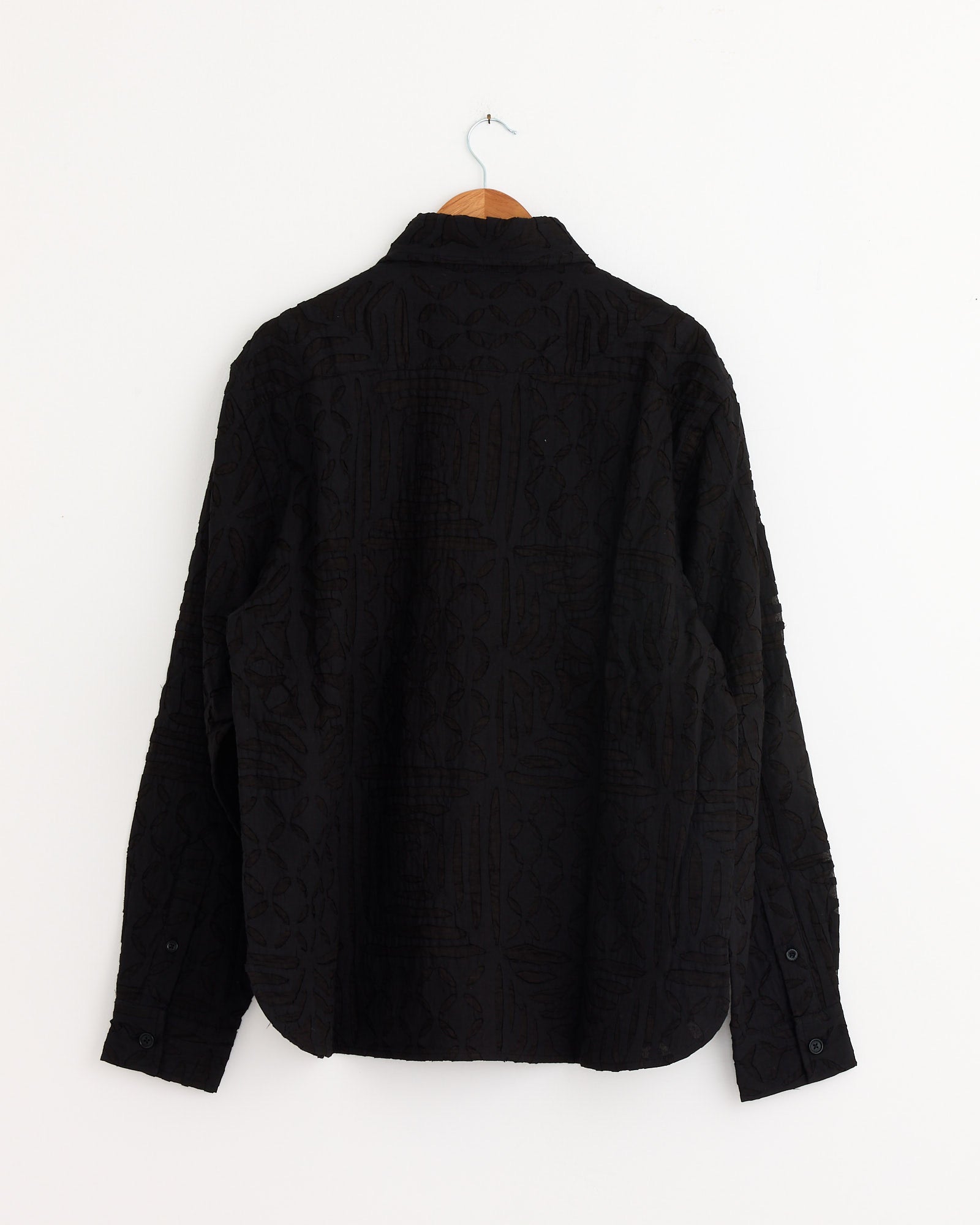The Luis Shirt in Black by Kardo—a long-sleeve, button-up khadi cotton shirt with a textured pattern—is shown from the back on a wooden hanger against a plain white wall.