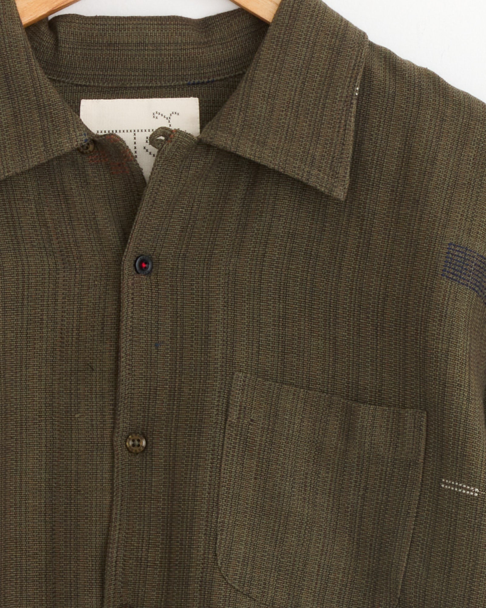 A close-up of the Kardo Chintan Shirt in Olive, a boxy-fit, striped, naturally dyed cotton shirt, showcases the collar, one brown button, chest pocket, and a partial inner label on a wooden hanger.