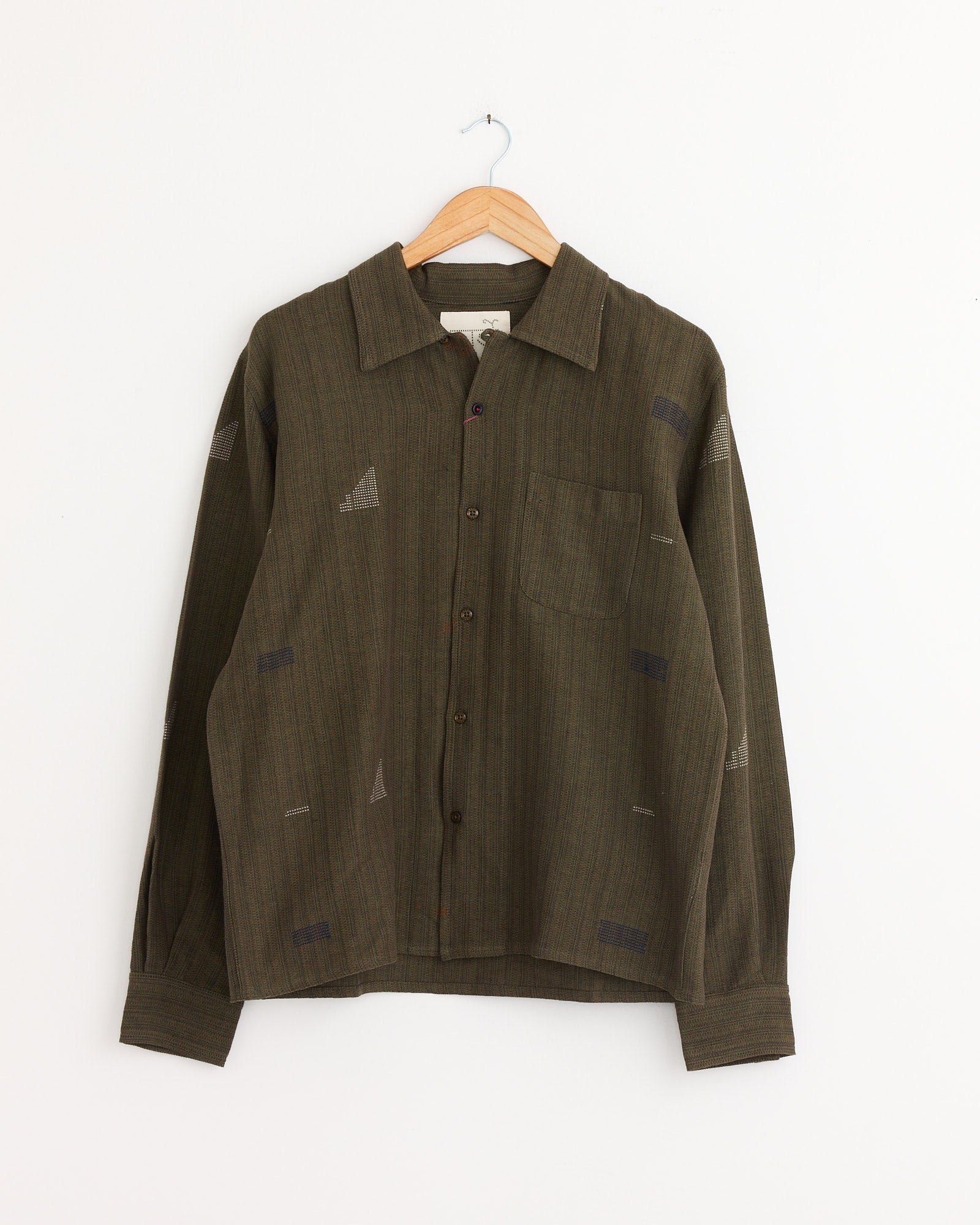 The Kardo Chintan Shirt in Olive is a boxy-fit, long-sleeve shirt crafted from naturally dyed cotton with subtle geometric patterns, a collar, and chest pocket, shown hanging on a wooden hanger against a white background.