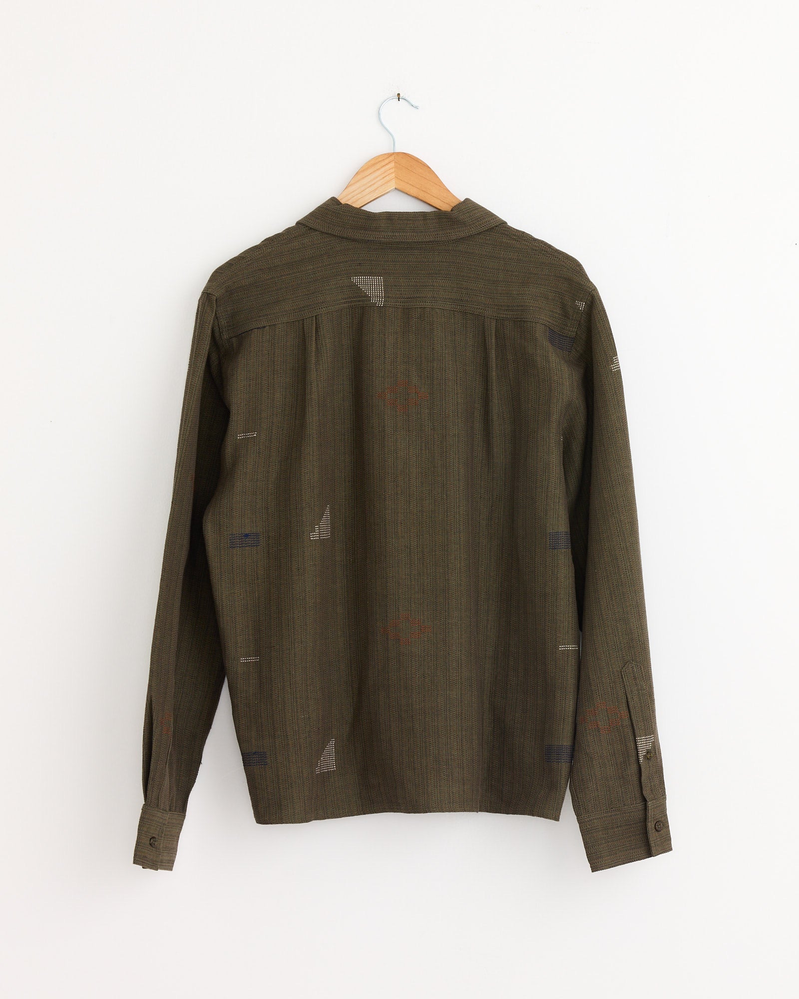 The Chintan Shirt in Olive by Kardo, a boxy-fit long-sleeve shirt with a subtle pattern, made from naturally dyed cotton, is shown from the back on a hanger against a plain white background.