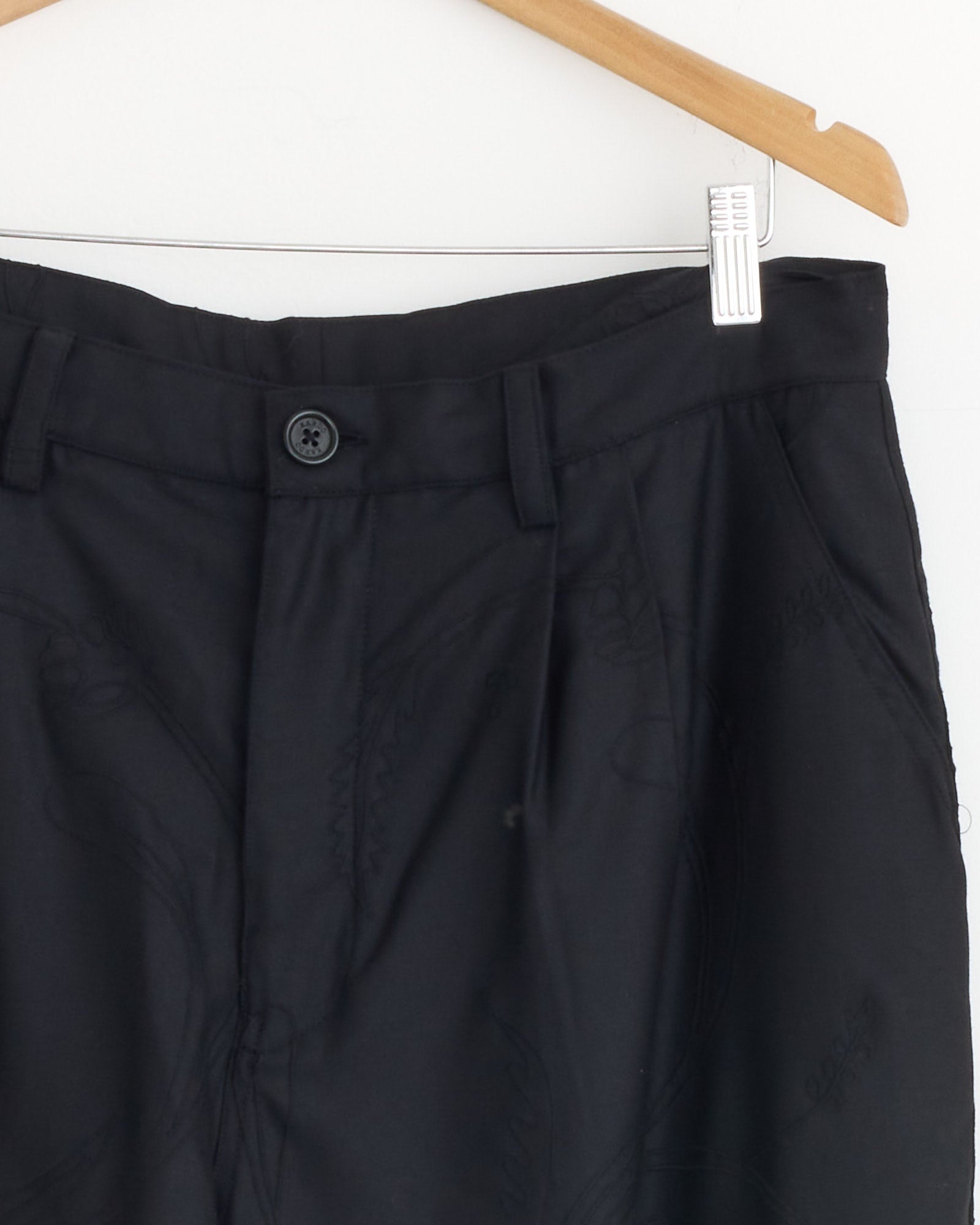 Close-up of the Kardo Antonio 2.0 Trouser in Black hanging on a wooden hanger against a white wall, highlighting the waistband, button, and front pleats.