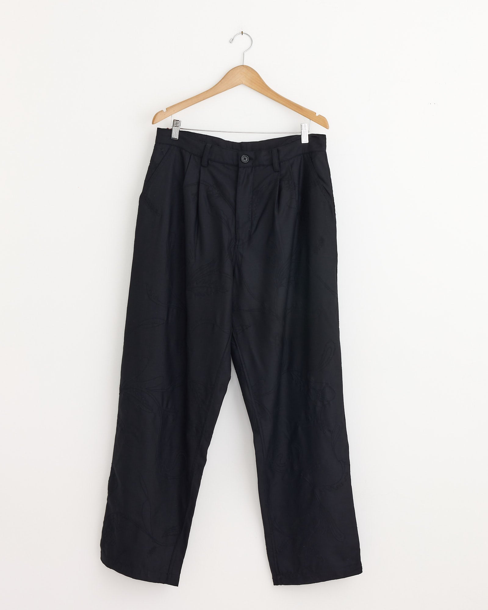 The Antonio 2.0 Trouser in Black by Kardo, crafted from handloom cotton, hangs on a wooden hanger against a white wall. These loose-fitting pants feature a button and zipper closure with subtle seam details.