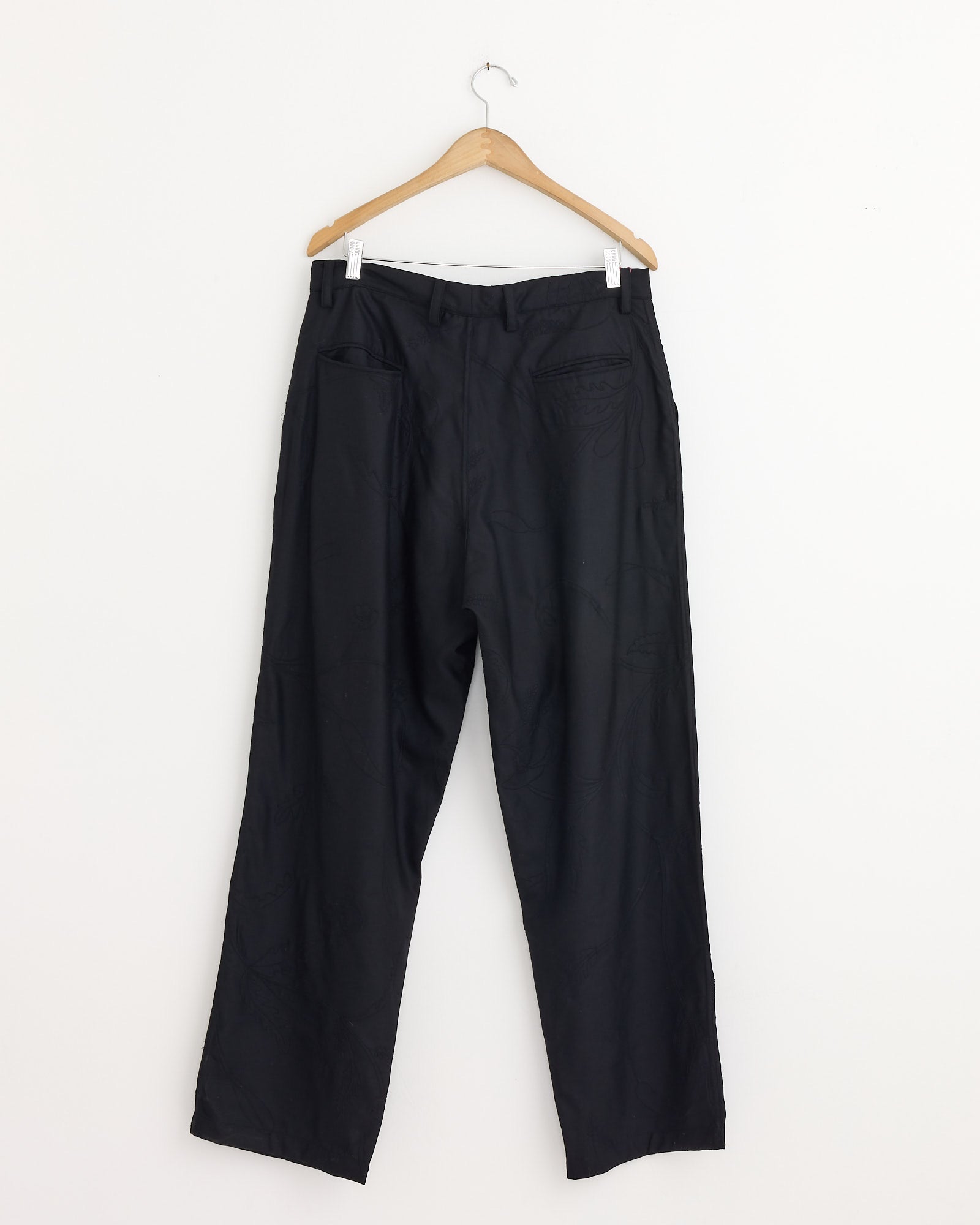 The Kardo Antonio 2.0 Trouser in Black, featuring a straight-leg fit and double pleats, hangs on a wooden hanger against a plain white wall.