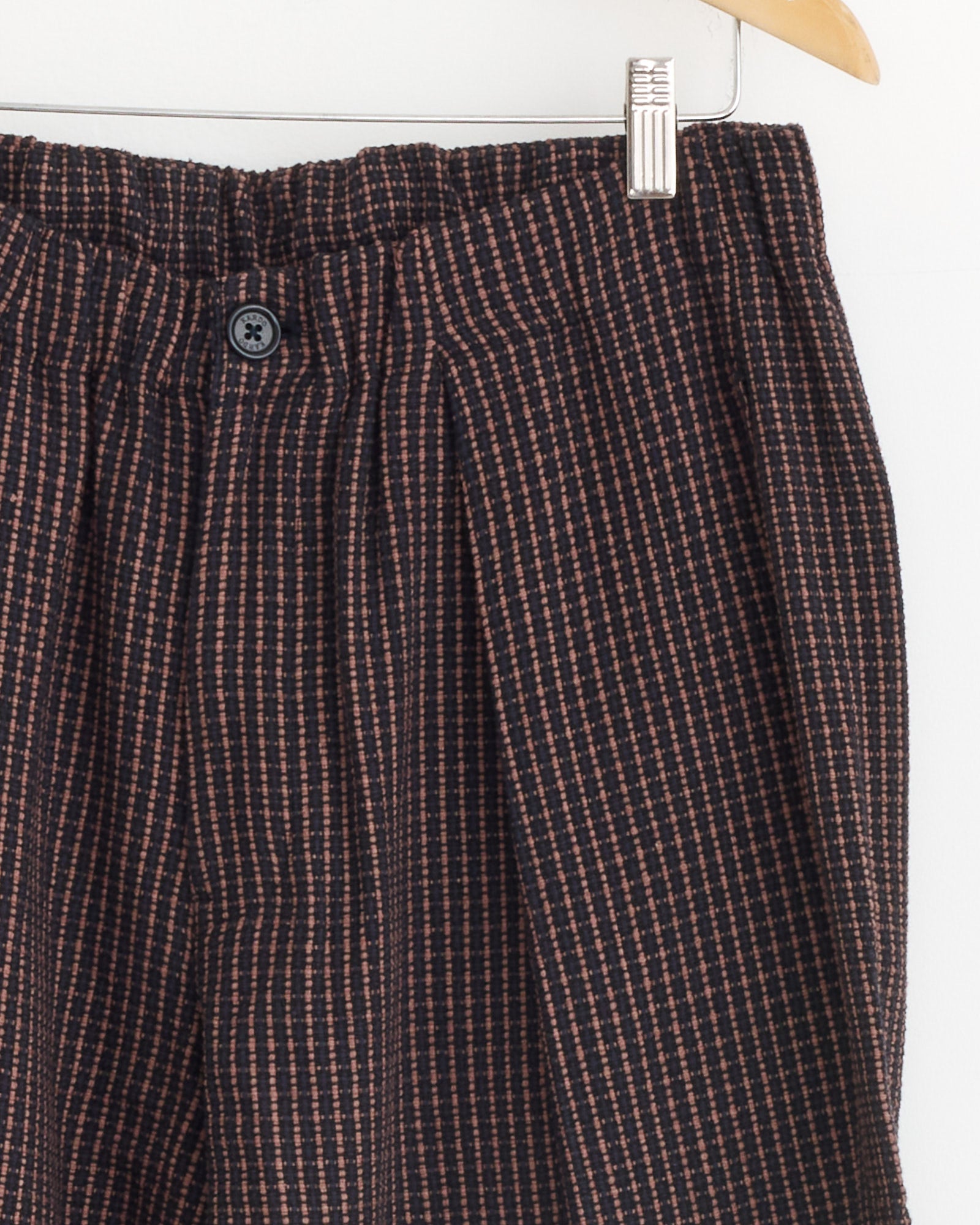 Close-up of the Kardo Taka Trouser in Ganache, a relaxed fit corduroy style with an elastic waist and button, showcasing dark fabric with a pink grid pattern, displayed on a wooden hanger against a white background.