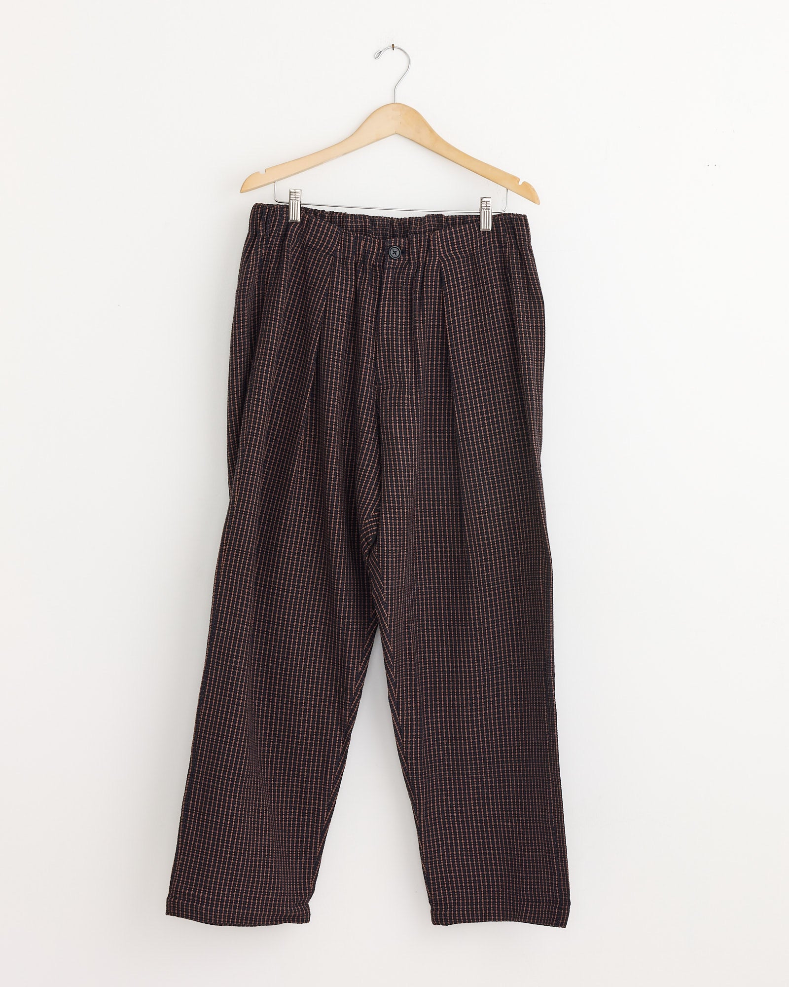 The Kardo Taka Trouser in Ganache, featuring a relaxed fit, dark plaid design, elastic waistband, and button closure, hangs on a wooden hanger against a plain white wall.