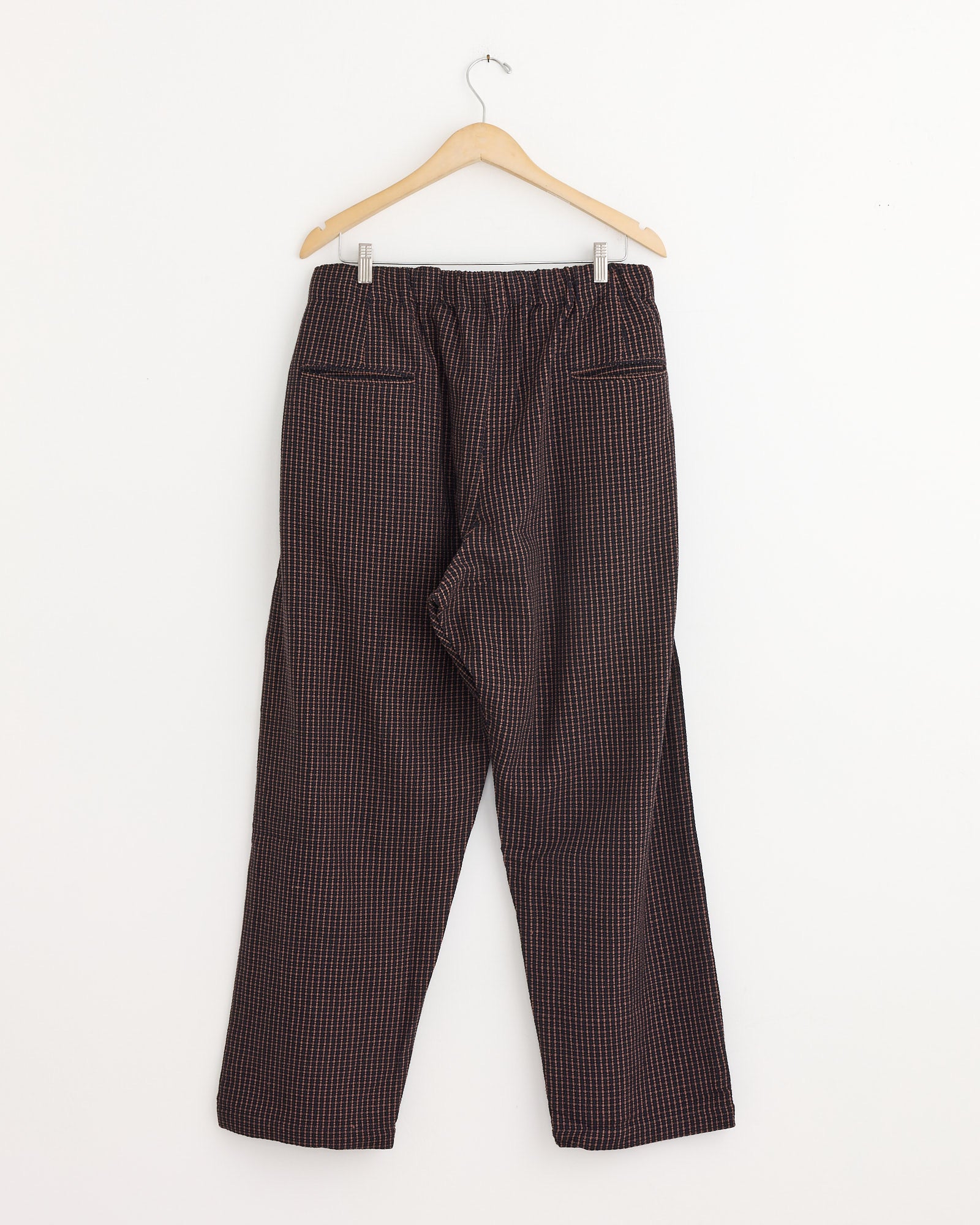 The Taka Trouser in Ganache by Kardo, featuring a high-waisted, relaxed fit and straight-leg corduroy design with a black and red checkered pattern, hangs on a wooden hanger against a plain white wall.