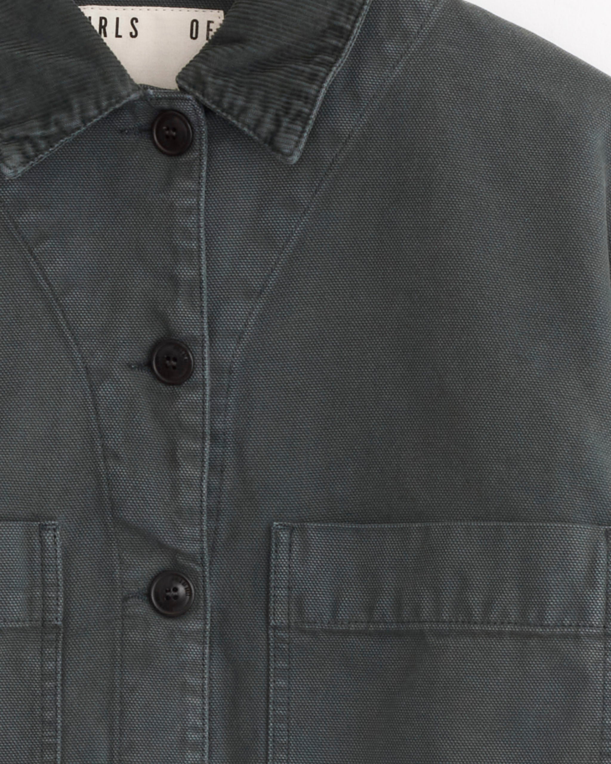 Close-up of the Girls of Dust Woodsy Jacket in forest green, featuring visible stitching, a collar, black buttons, and two chest pockets. A white label with black text is partially visible inside the collar.