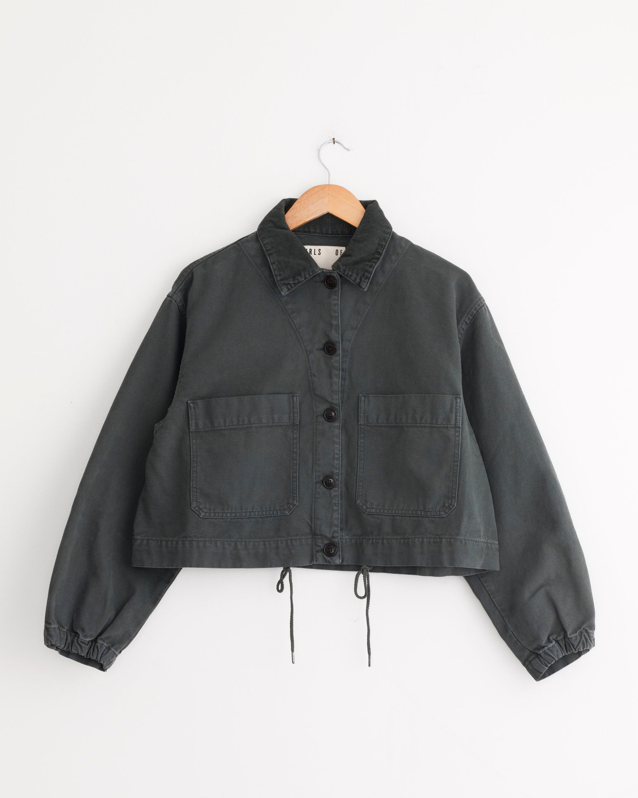 The Girls of Dust Woodsy Jacket in Forest Green, featuring a collar, button-down front, two large front pockets, and elastic cuffs, hangs on a wooden hanger against a white background.