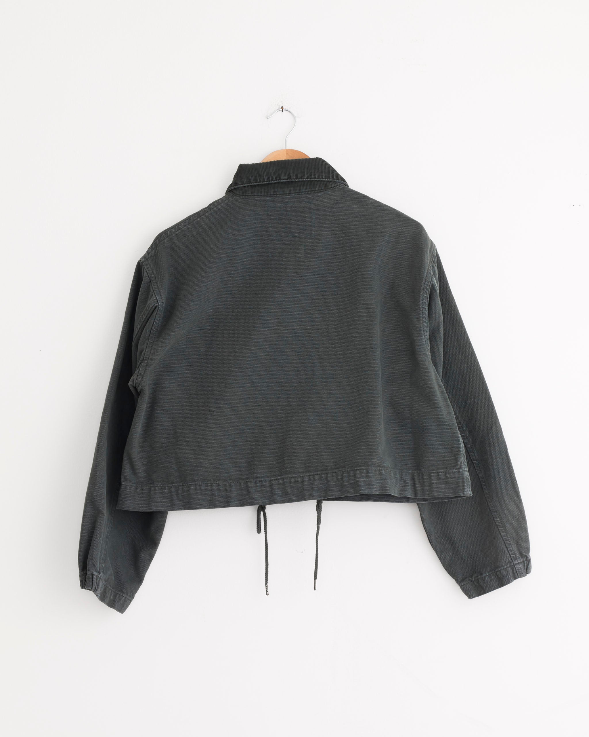 A forest green, long-sleeved Woodsy Jacket by Girls of Dust hangs on a wooden hanger against a plain white wall, shown from the back. This cropped worker jacket features a collar, drawstring hem, and relaxed fit.