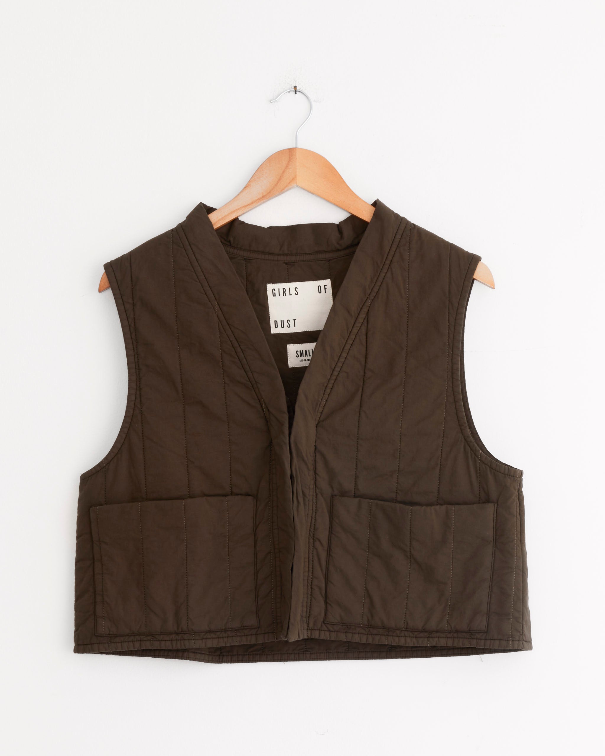 The Girls of Dust Cork Vest in Java Brown, a quilted cotton design with two large front pockets, hangs on a wooden hanger against a plain white wall. The inner label displays the brand name GIRLS OF DUST.