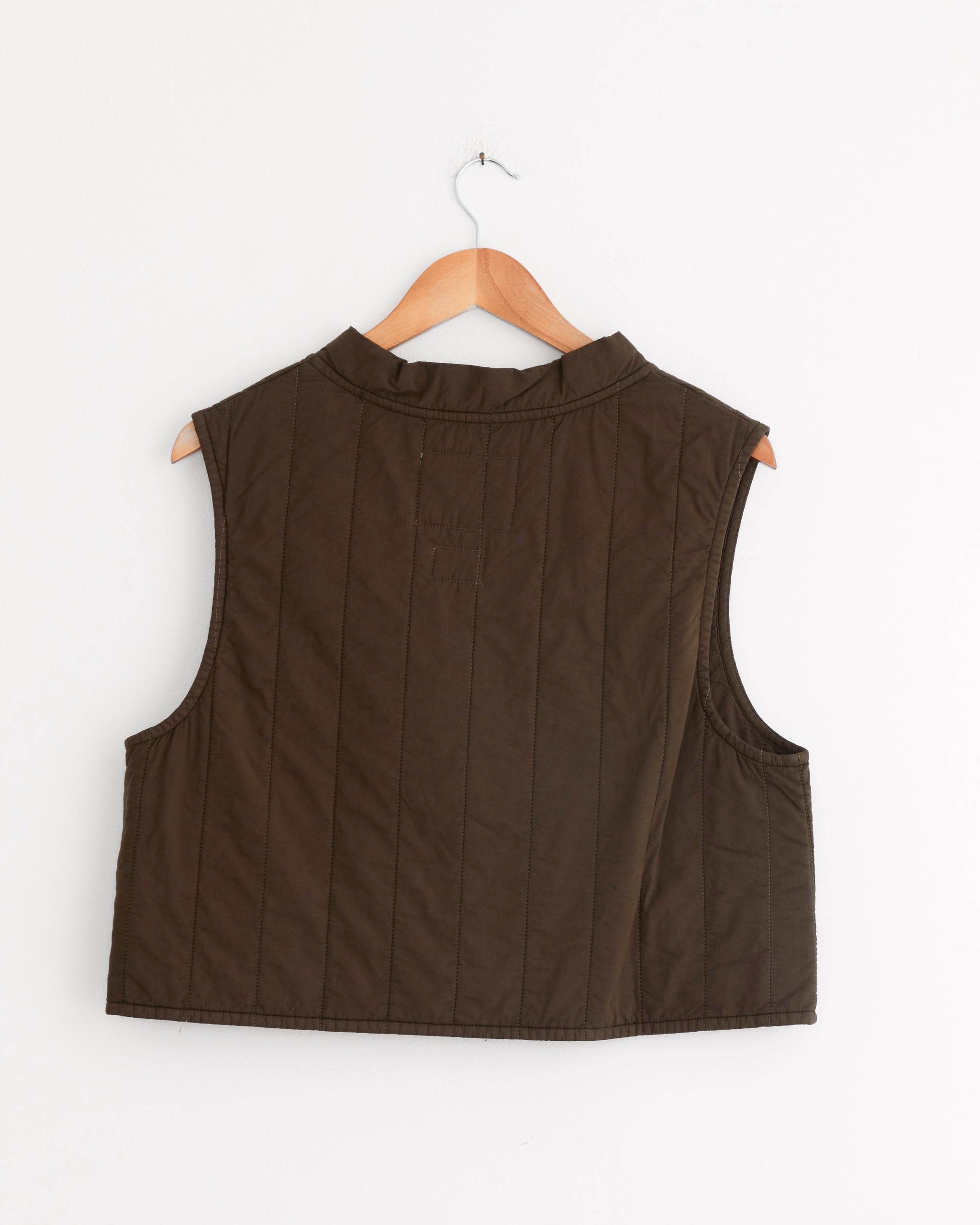 The Girls of Dust Cork Vest in Java Brown hangs on a wooden hanger against a white wall, displayed sleeveless and quilted with the back facing forward.