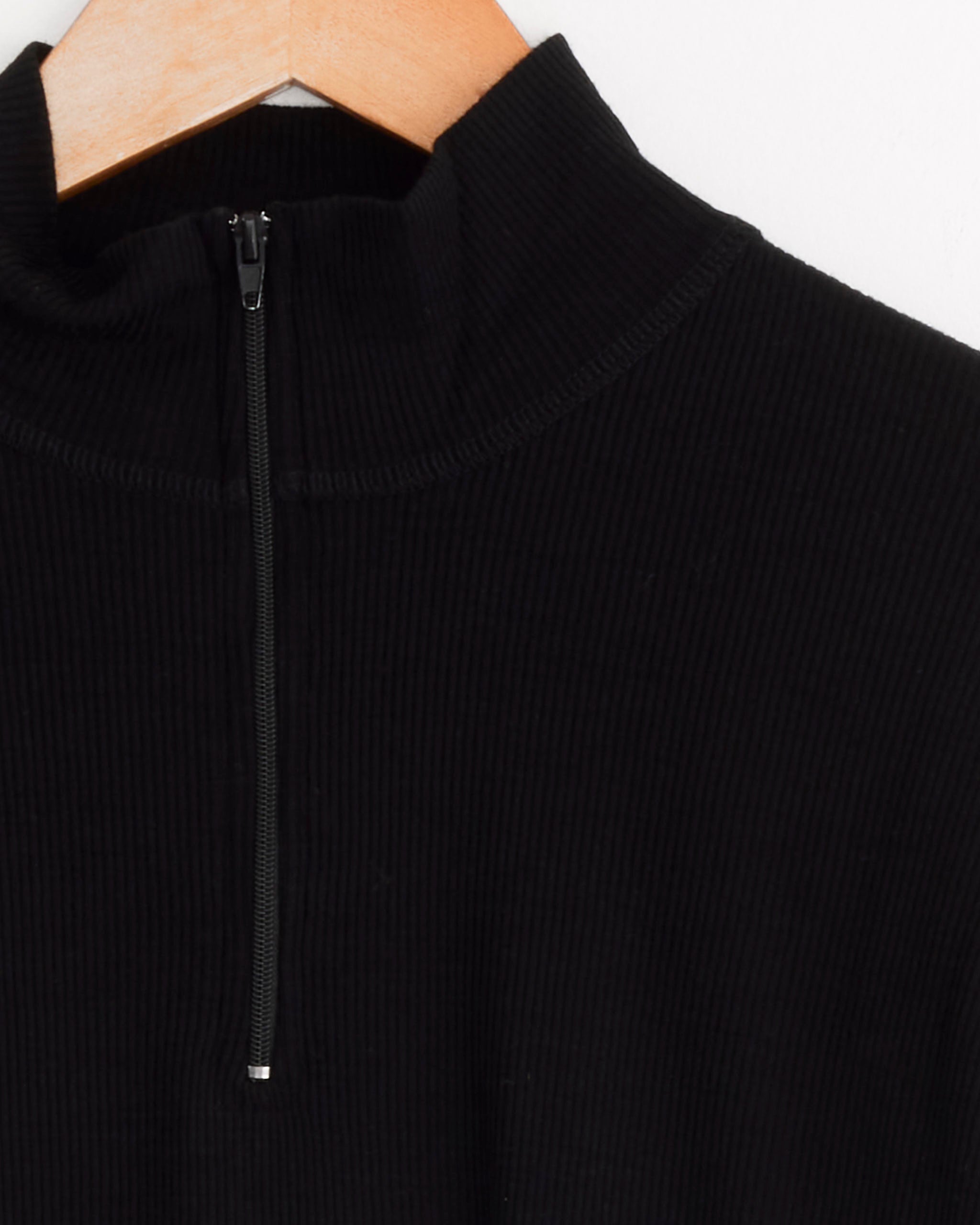 The Girls of Dust Zipper Pull Over in Black is a ribbed wool pullover with a high collar and partial front zipper, shown on a wooden hanger. It brings effortless sophistication to any wardrobe.