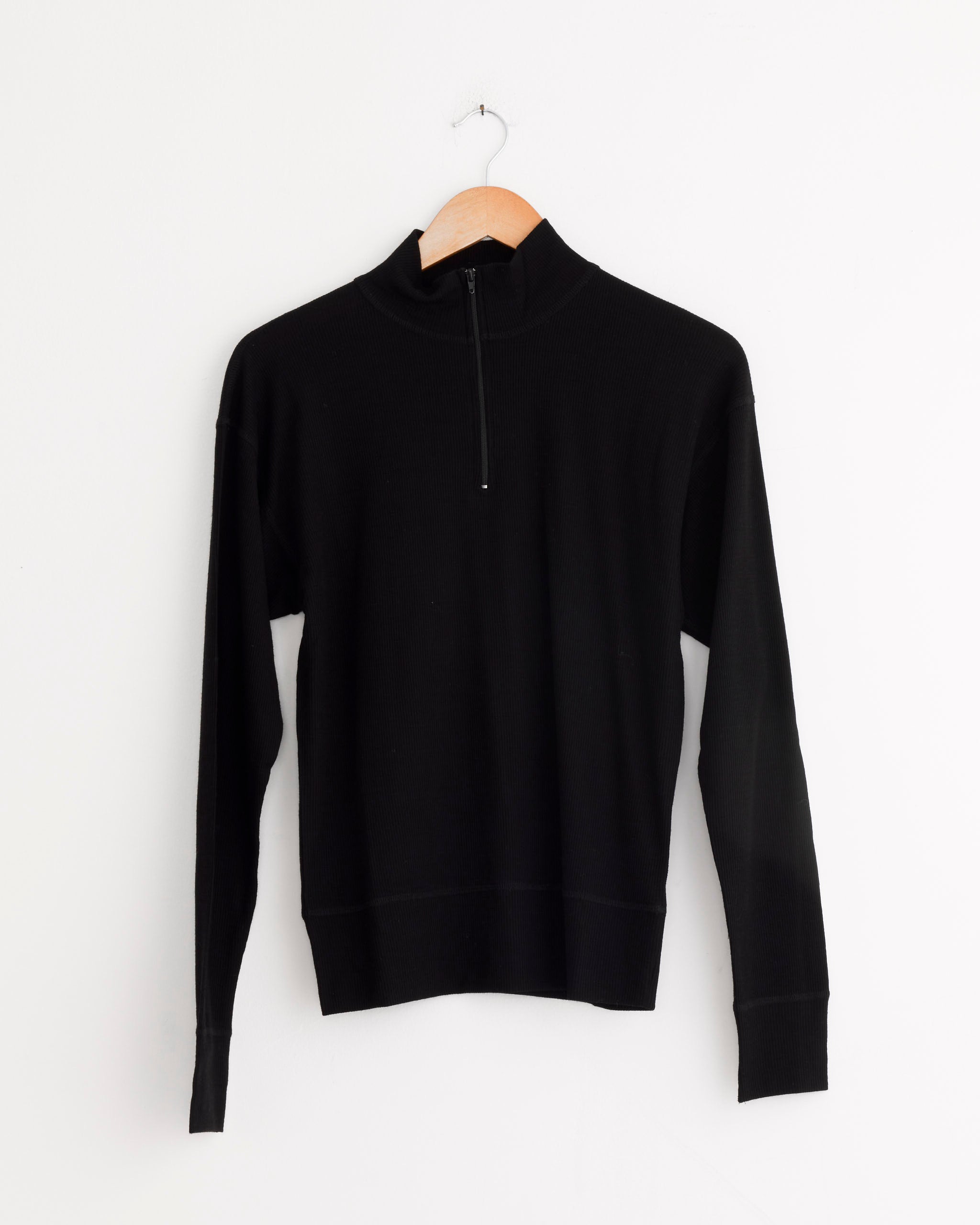 The Girls of Dust Zipper Pull Over in Black is a wool sweater with a stand-up collar and half-zip front, shown on a wooden hanger against a white background. Classic style meets cozy comfort.