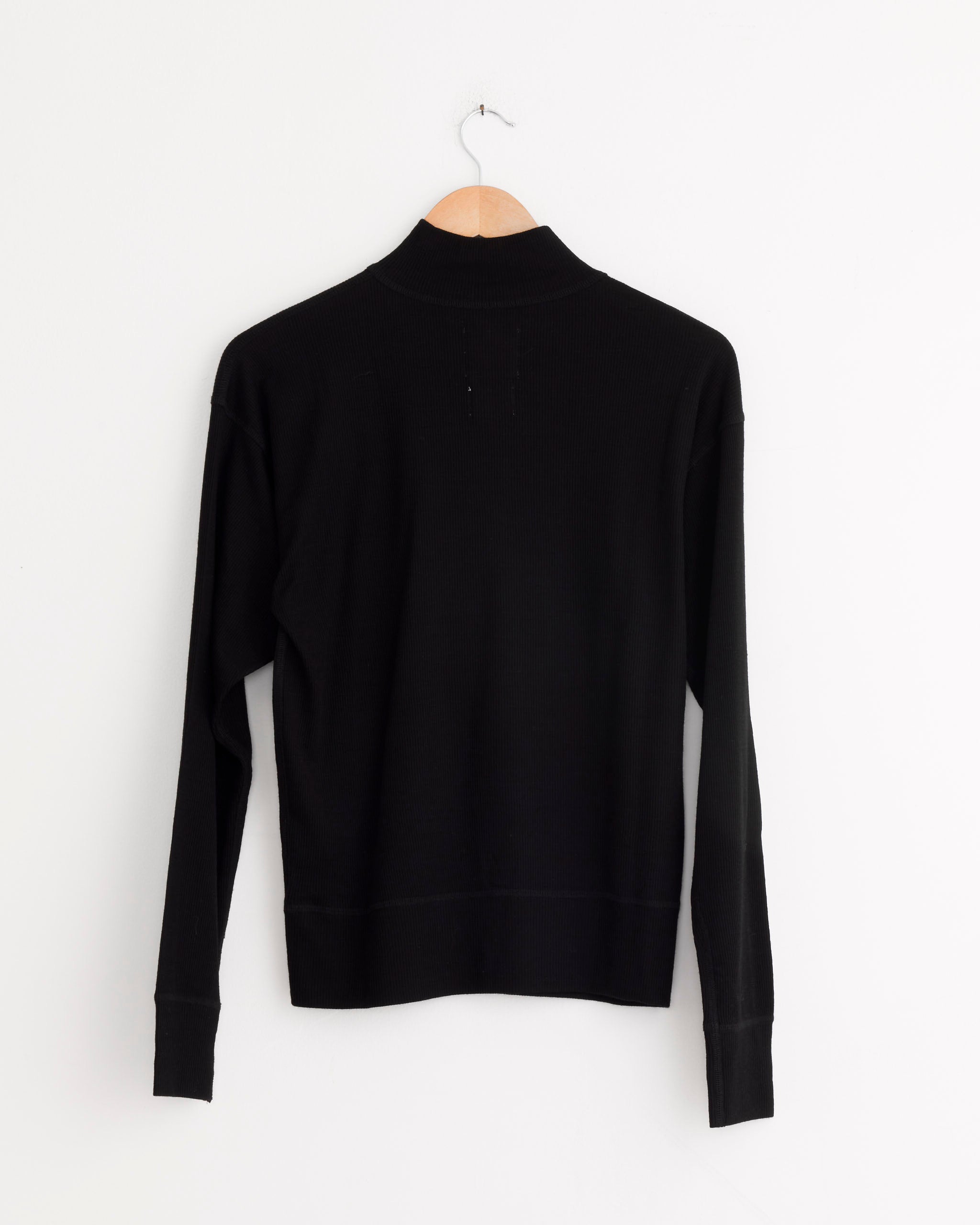 The Girls of Dust Zipper Pull Over in Black, a long-sleeve sweater, is displayed from the back on a wooden hanger against a plain white wall.