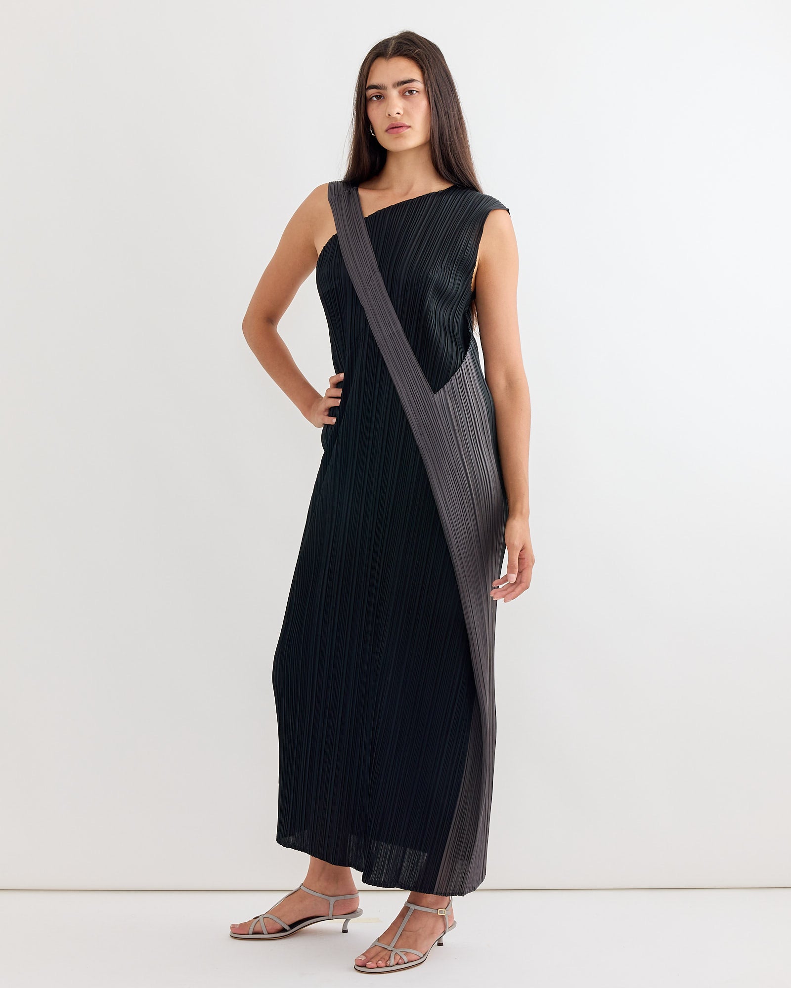 A woman wears the Pointe Dress in Black by Pleats Please Issey Miyake—an ankle-length, sleeveless style with vertical pleats and a bold grey diagonal panel. Need help with sizing or fit? Our online chat is here to assist you.