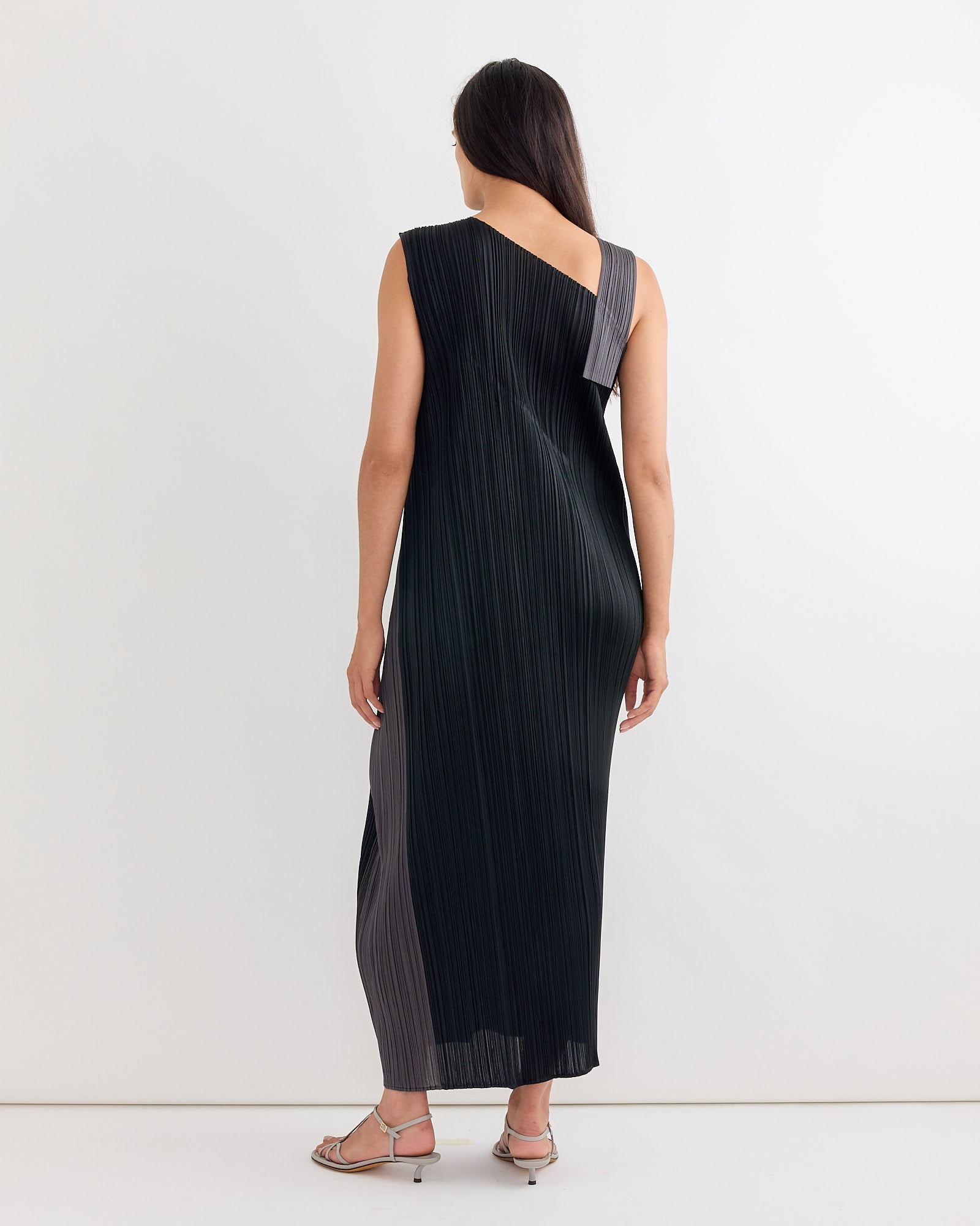 A woman faces away, wearing the Pleats Please Issey Miyake Pointe Dress in Black—a sleeveless, ankle-length pleated dress with an asymmetrical neckline and wide strap. She has long dark hair and light sandals. For sizing help, use our online chat. Plain white background.