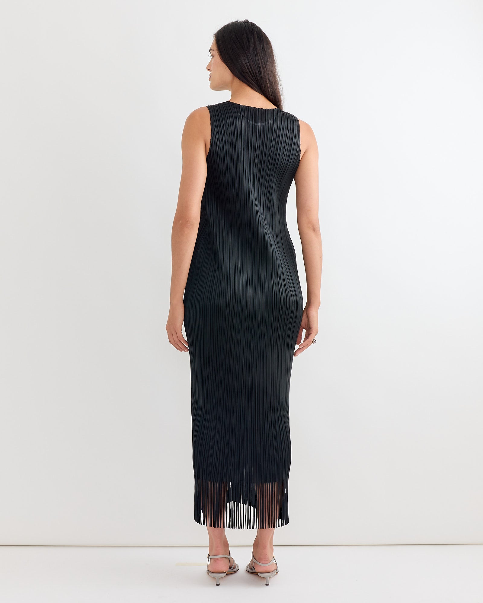 A woman with long dark hair models the Wave Dress in Black by Pleats Please Issey Miyake. She stands with her back to the camera, highlighting the ankle-length pleated silhouette and fringe hem against a plain white background.