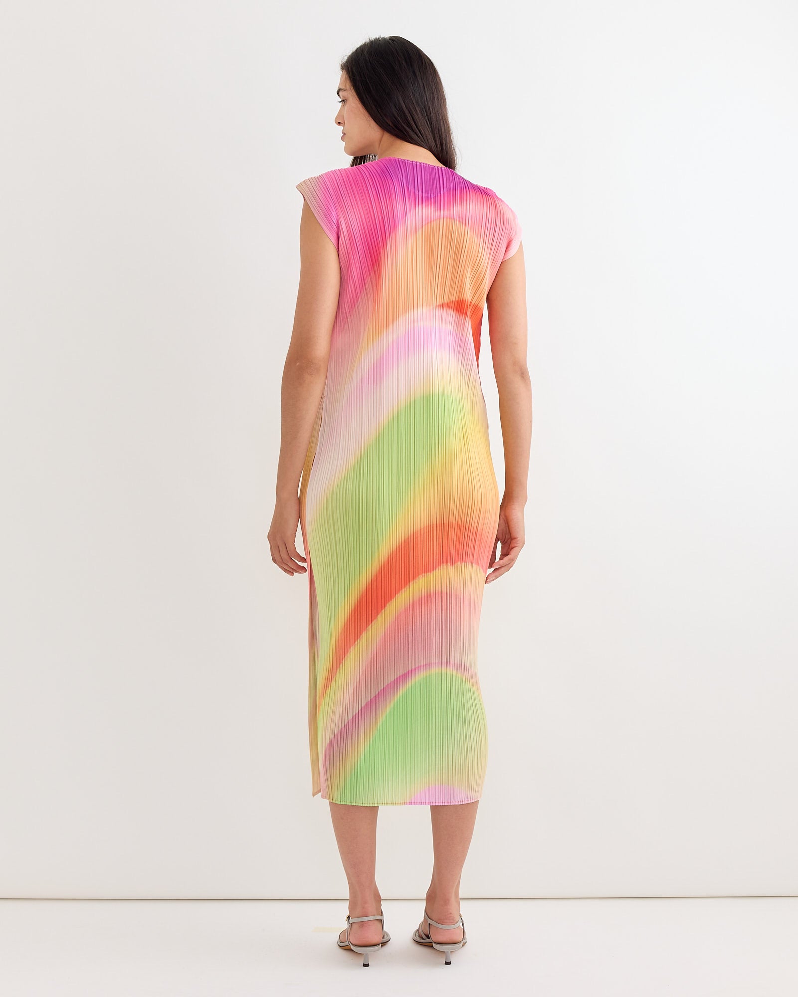 A woman with straight dark hair and heeled sandals faces away, wearing the Pleats Please Issey Miyake Groove Dress in Bright Pink—a sleeveless, calf-length piece with signature pleats and a pastel gradation print.