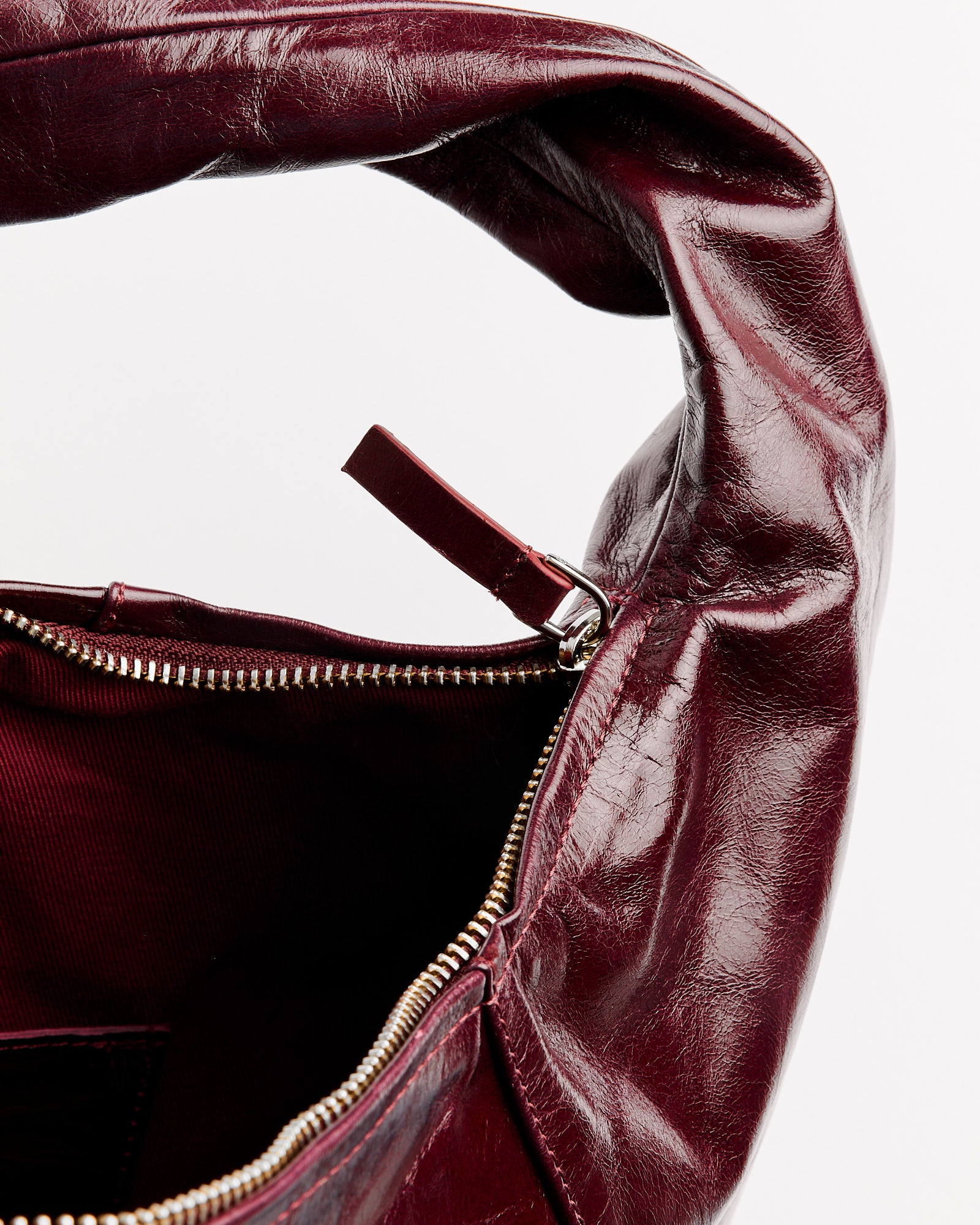Close-up of the top of the Gimaguas Paula Bag in Burgundy, featuring shiny, slightly wrinkled leather with an open zipper and a glimpse of the dark fabric interior. Contact our online chat for sizing or fit questions.