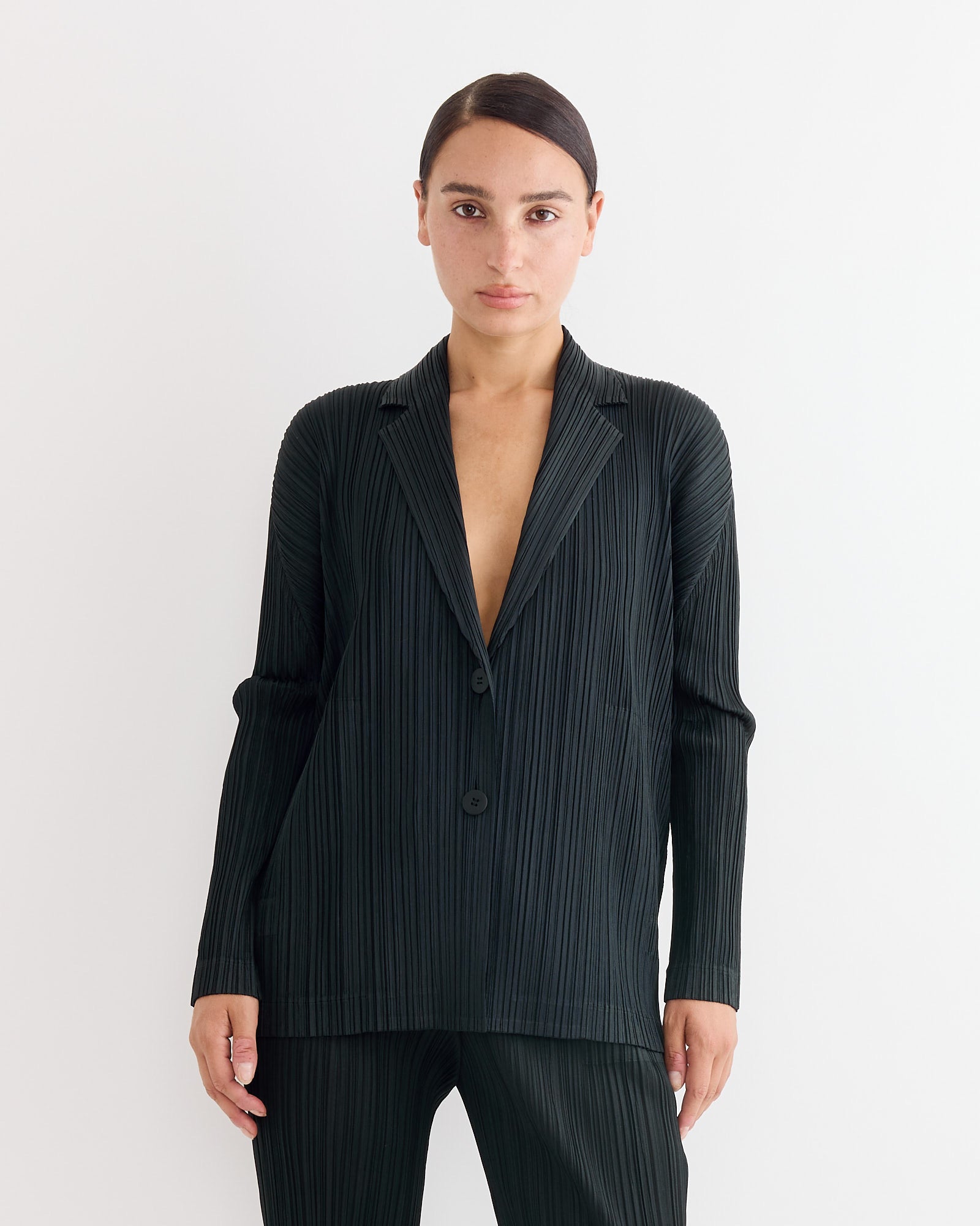 A person with dark hair pulled back models the Pleats Please Issey Miyake Jacket in Black, featuring pleats, buttons, and a sleek fit, paired with matching pants against a plain white background.