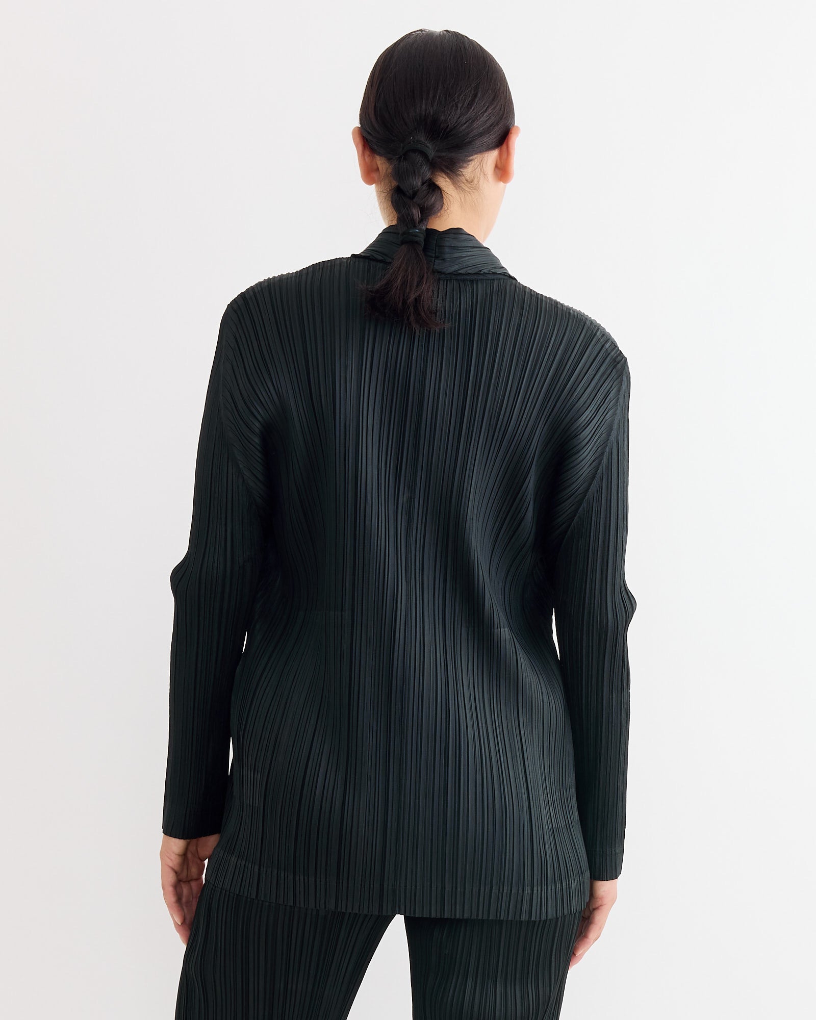 A person with dark hair in a low braid stands with their back to the camera, wearing the Pleats Please Issey Miyake Jacket in Black—ideal for anyone seeking the perfect fit or sizing advice.