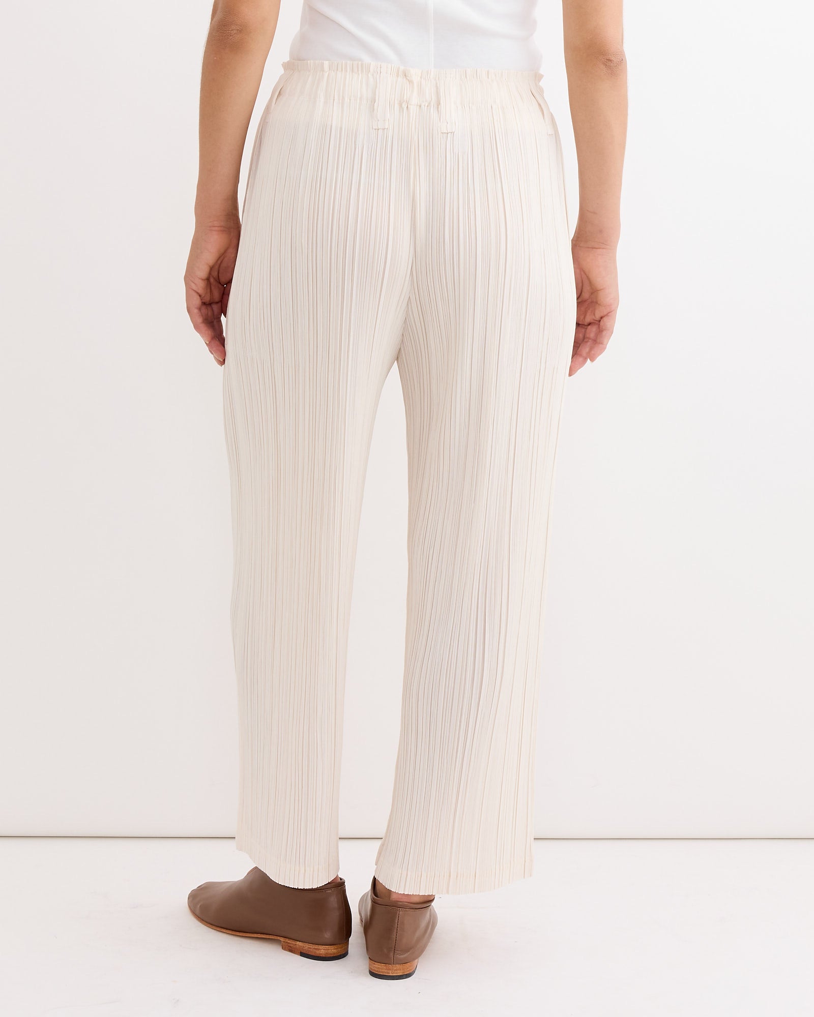 A person stands facing away, wearing Pleats Please Issey Miyake Thicker Bottoms 2 Pant in Off White with brown slip-on shoes; the background is plain white, and only part of their upper body is visible.