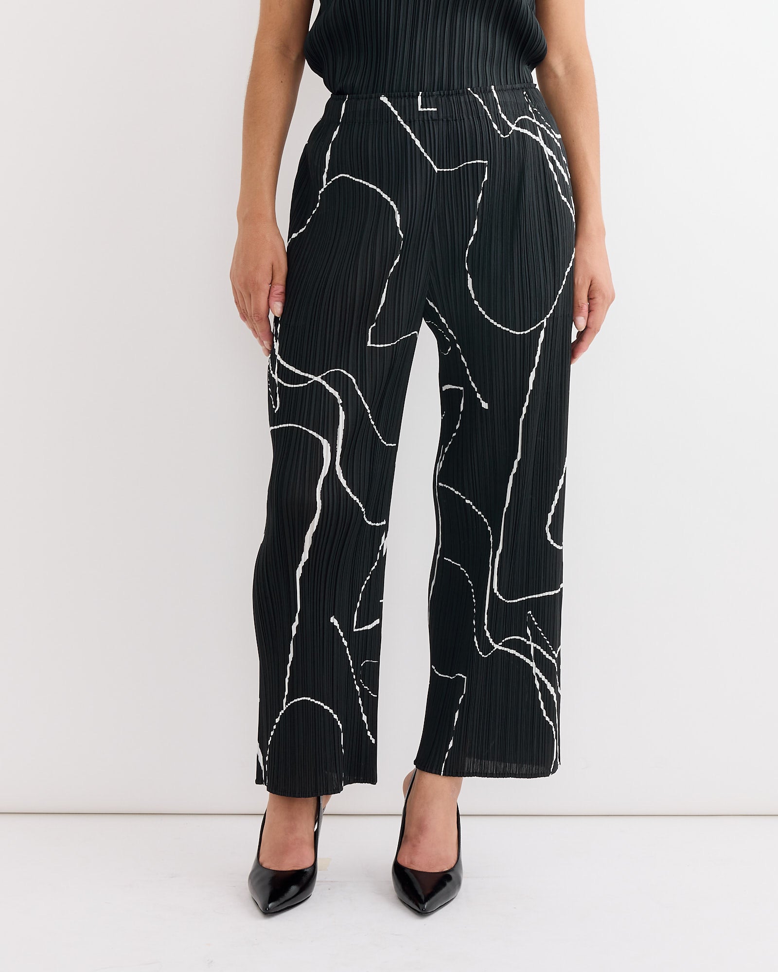A person stands against a plain white background wearing the Pleats Please Issey Miyake Pause Pant in Black featuring white abstract line patterns, styled with black high heels.