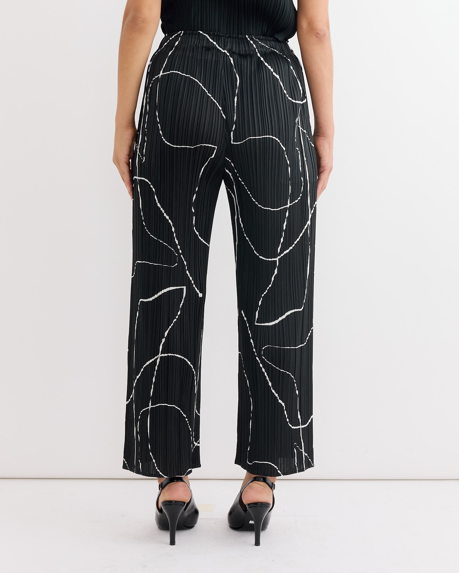 A person wears Pleats Please Issey Miyake's Pause Pant in Black, featuring an abstract white line pattern and straight-leg fit, styled with black heels for a sleek, modern look against a white background.